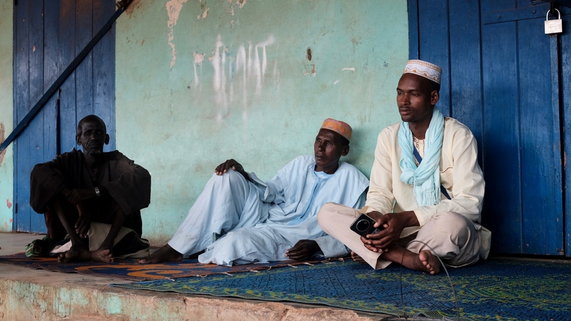 Cette image montre trois hommes assis sur un tapis, à l'extérieur d'un bâtiment aux murs de couleur turquoise. L'atmosphère semble calme et détendue. Ils portent des vêtements traditionnels : deux d'entre eux sont vêtus de tuniques, tandis que le troisième porte un chapeau. L'un des hommes est assis en tailleur, tandis que les deux autres sont en position plus détendue, le dos appuyé contre le mur. À côté d'eux, il y a un petit seau en métal d'où s'écoule un peu d'eau. En arrière-plan, des murs sont partiellement recouverts de graffiti. L'environnement semble rural, avec une lumière douce qui illumine la scène.