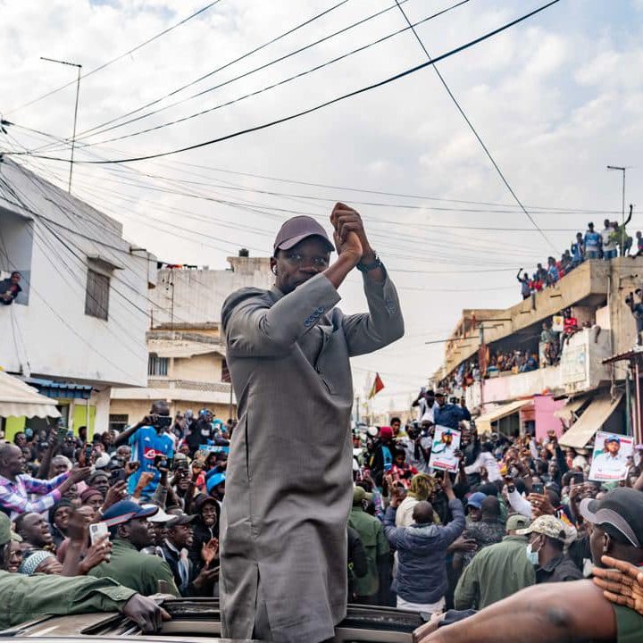 L'image montre une scène dynamique et animée dans une rue bondée. Au centre, un homme, vêtu d'une tenue grise, se tient debout sur un véhicule, levant les bras avec enthousiasme. Les gens autour de lui, une foule importante, applaudissent et expriment leur soutien. On peut entendre des cris de joie et des chants de la part des personnes qui entourent le véhicule. À l'arrière-plan, des bâtiments et des personnes sur les balcons ajoutent à l'atmosphère de ferveur et de célébration. Le ciel est partiellement nuageux, créant un éclairage intéressant sur cette scène vibrante.
