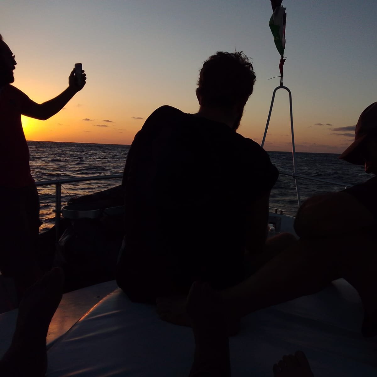 The image shows a group of people on a boat at sunset. The sky is painted in warm hues of orange and yellow as the sun sets on the horizon. Two people are visible, sitting with their backs to the camera, seemingly enjoying the view. One person is standing and holding a phone, possibly taking a photo. The water appears calm, reflecting the colors of the sky. The overall atmosphere is serene and relaxed.
