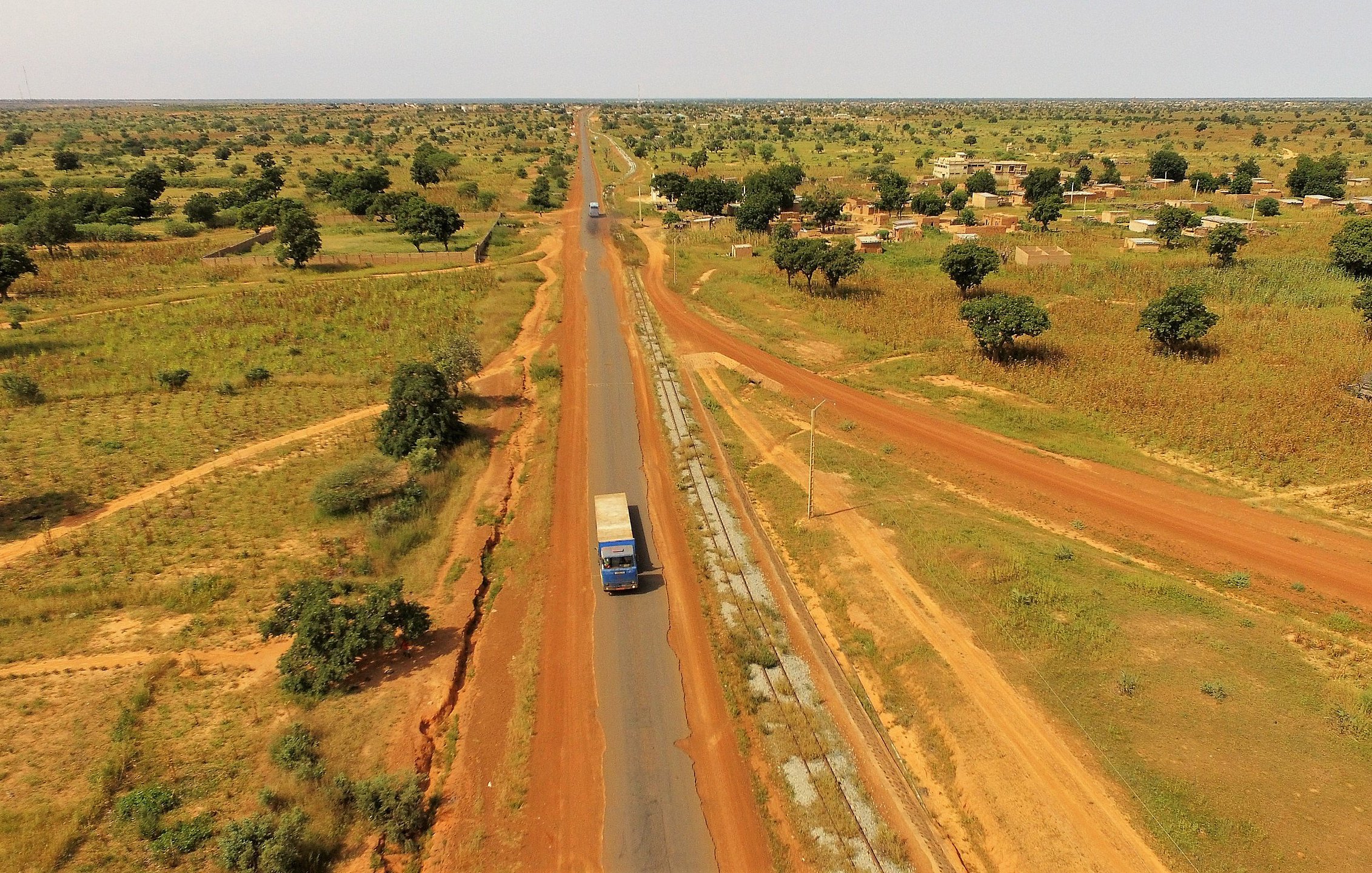 Cette image présente un paysage rural étendu, où une route goudronnée traverse une vaste plaine. Sur la droite, on aperçoit un camion bleu qui circule, indiquant une activité de transport. De part et d'autre de la route, le sol est d'un rouge terreux, typique de certaines régions africaines, semé de petites collines et de végétation éparse, notamment des arbres et des buissons. Sur la gauche de l'image, on peut distinguer des habitations simples, suggérant la présence d'un village. L'arrière-plan est composé de champs et d'arbres, renforçant l'impression de tranquillité et d'espace ouvert. L'ensemble évoque une atmosphère de ruralité, avec une nature généreuse, baignée par une lumière intense.