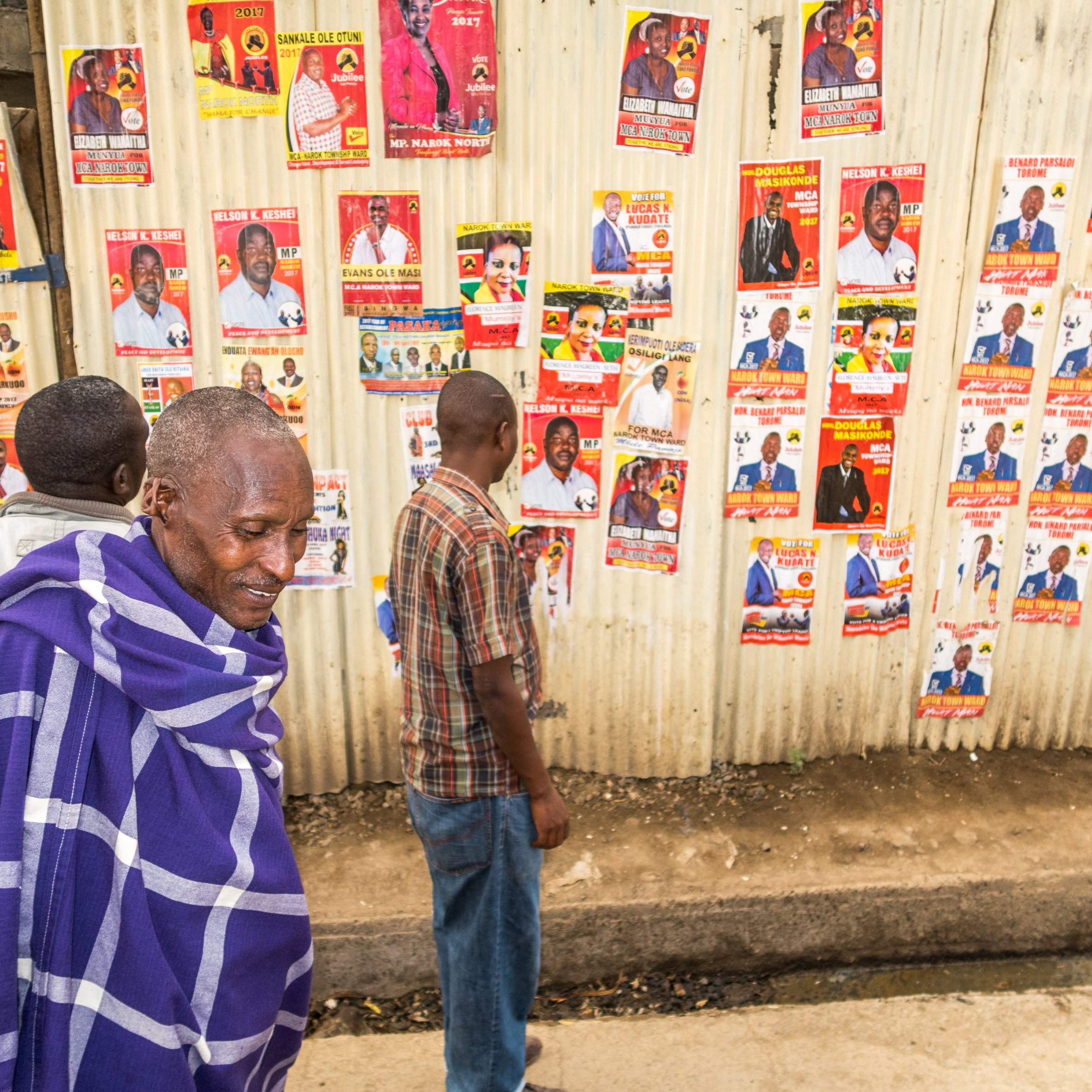 L'image montre un groupe de personnes marchant le long d'un mur recouvert de nombreuses affiches. Ce mur en tôle est tapissé de posters colorés présentant des candidats politiques, avec des visages souriants et des informations écrites. Au premier plan, un homme est vêtu d'un châle traditionnel bleu et blanc, semblant contempler les affiches. À ses côtés, d'autres personnes se déplacent lentement, examinant les posters tandis que le sol est pavé, dans un environnement urbain. L'atmosphère semble à la fois animée et réfléchie, avec un mélange de culture locale et d'engagement politique.