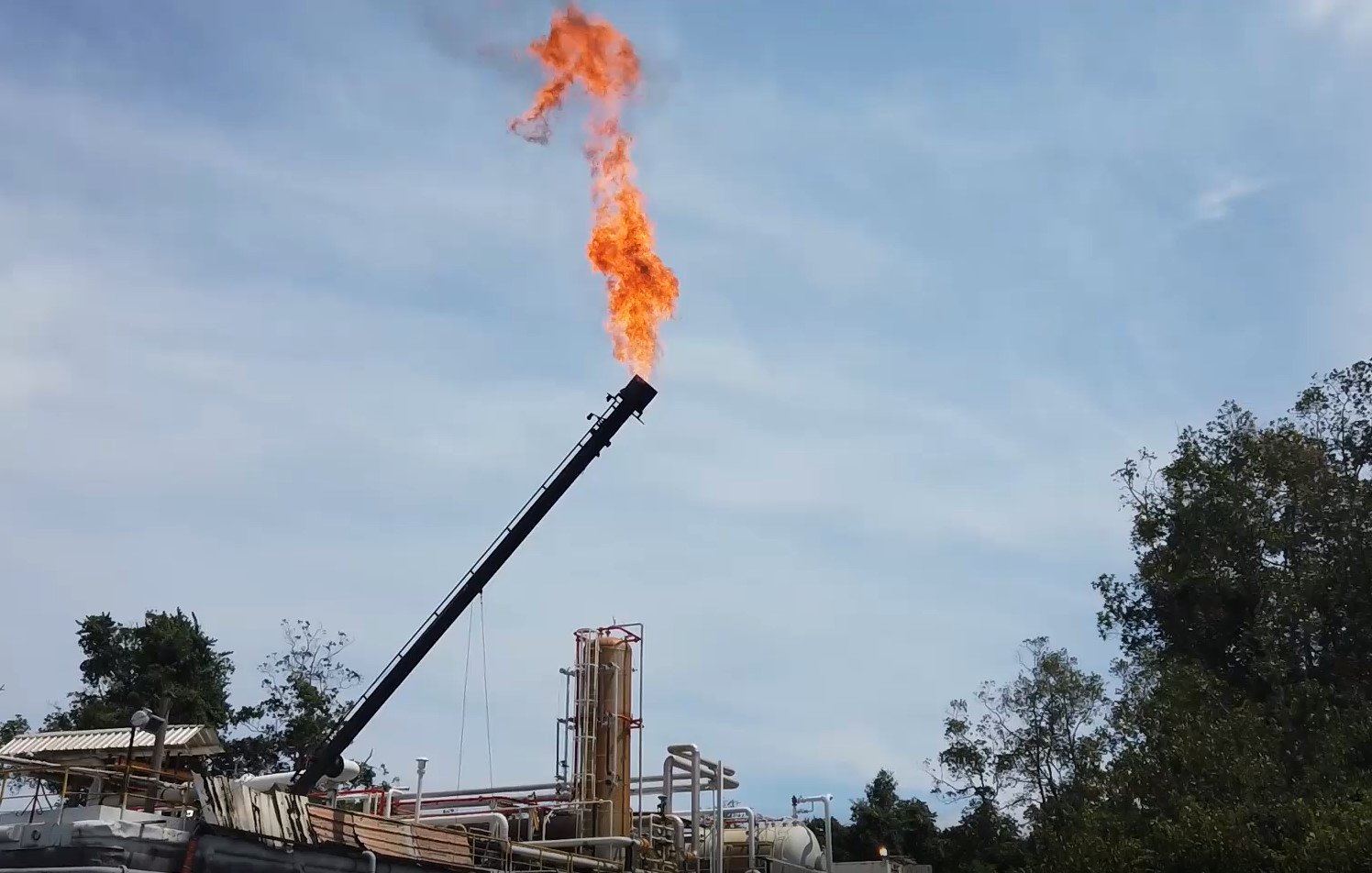 L'image présente une installation industrielle en plein air, probablement liée à l'extraction de pétrole ou de gaz. Au centre, un tube long et noir s'élève vers le ciel, d'où s'échappe une flamme vive et orange, créant un fort contraste avec le ciel bleu clair en arrière-plan. On aperçoit également des structures en métal, comme des réservoirs et des tuyaux, sur le site. De la végétation verte entoure l'installation, ajoutant une touche naturelle à cette scène industrielle. Le tout évoque une activité intense et potentiellement bruyante.