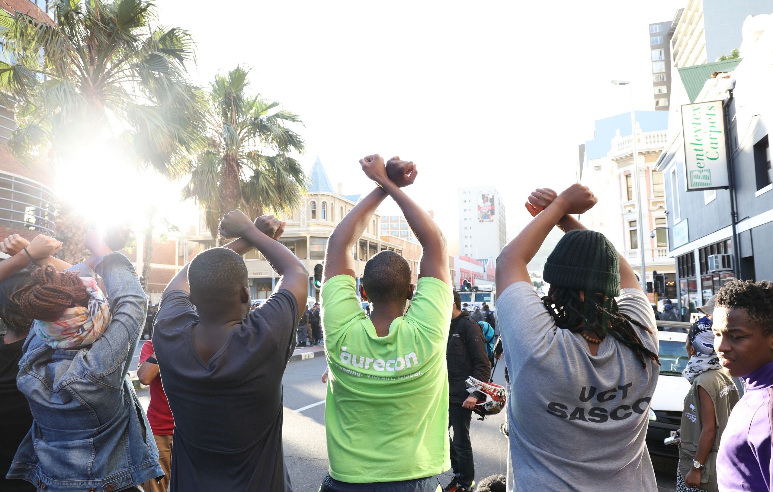 L'image montre un groupe de personnes vues de dos, levant les bras au-dessus de leurs têtes, formant un geste de solidarité ou de protestation. Ils sont sur une rue animée, entourés de bâtiments urbains. On peut apercevoir des palmiers à l'arrière-plan, ce qui ajoute une note de chaleur à l'environnement. La lumière du soleil éclaire la scène, créant une ambiance dynamique. Les personnes portent des t-shirts de différentes couleurs, dont l'un est vert vif. L'ensemble dégage une impression de union et d'engagement collectif.