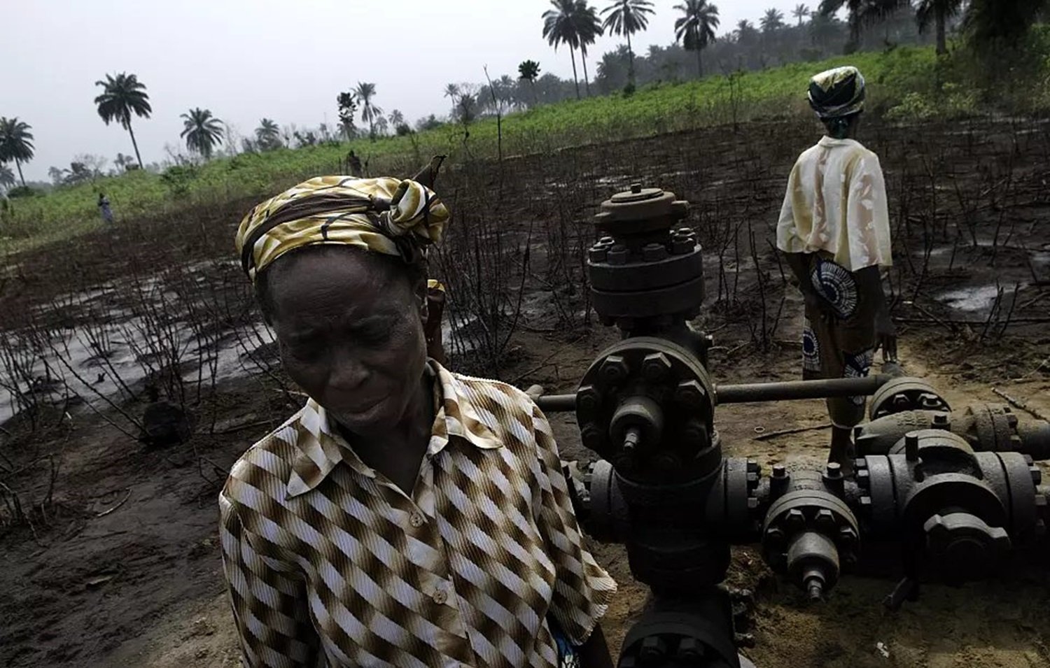 L'image montre un paysage rural où se trouvent des plantations de palmiers en arrière-plan. Au premier plan, une femme d'âge mûr, vêtue d'une blouse rayée et portant un foulard coloré, se tient près d'une infrastructure de forage pétrolier. Son expression est sérieuse et préoccupée. À l'arrière-plan, une autre personne, également en tenue décontractée, semble s'occuper de l'équipement. Le sol est noirci, suggérant une dégradation environnementale. L'atmosphère générale évoque une lutte pour préserver leur lien avec la terre face aux impacts de l'exploitation pétrolière.
