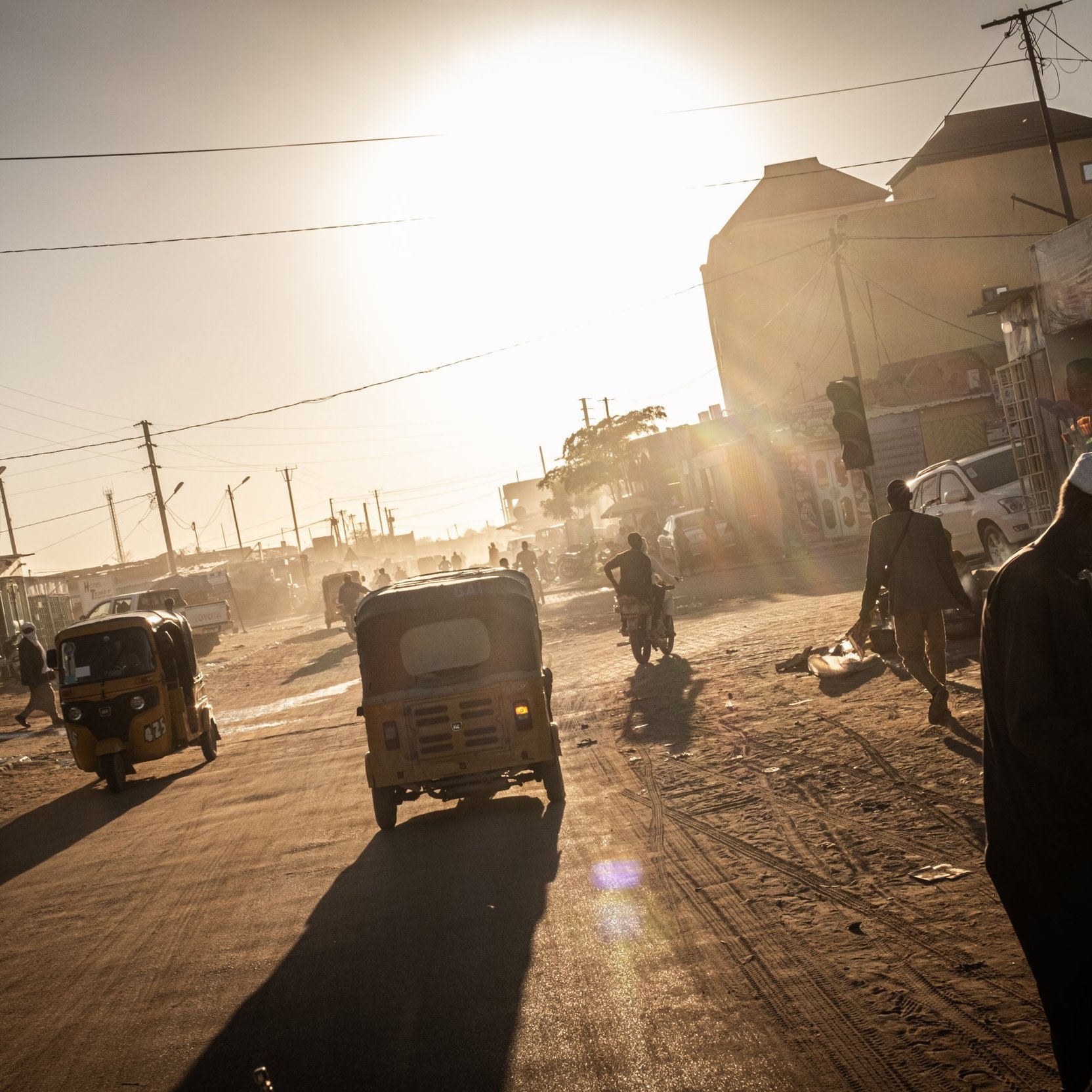 The image depicts a quiet, sandy street at sunset, where the golden light of the sun is visible on the horizon. On one side, there are simple, earth-toned walls of buildings, casting long shadows in the warm light. A few individuals are scattered along the street, with one person sitting and another standing near a donkey. The atmosphere suggests a peaceful, rural environment, with minimal traffic and a sense of tranquility as day transitions to night.
