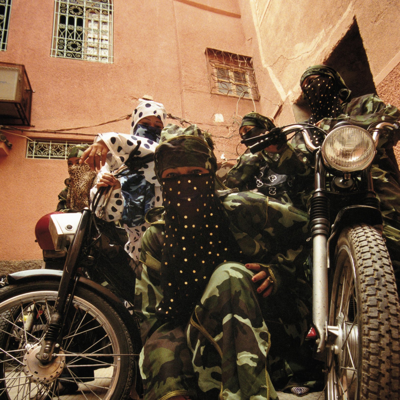 L'image montre un groupe de personnes habillées de manière distinctive, portant des vêtements camouflés, ainsi que des masques et des foulards ornés de paillettes ou de motifs. Ils se trouvent dans une rue d'une vieille ville, probablement dans un environnement urbain. À côté d'eux, deux motos ajoutent un élément de modernité à la scène. Leurs poses évoquent une certaine solidarité et un esprit de camaraderie. L'éclairage est naturel, suggérant un moment de la journée typique dans une ville animée. Les textures des vêtements contrastent avec le pavé de la rue, créant une ambiance unique.