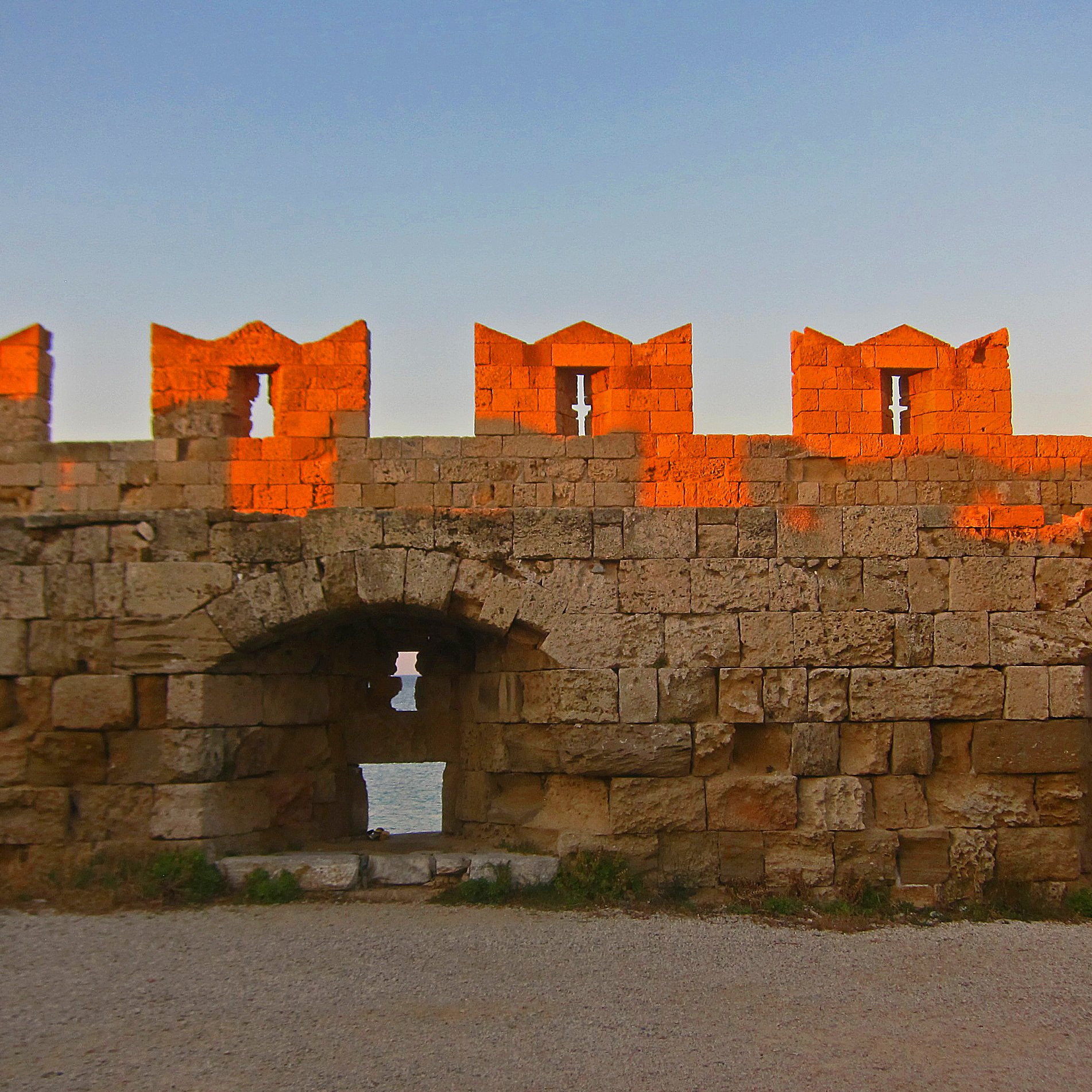 L'image montre un mur de fortification ancien, construit en pierres de taille. La lumière dorée du soleil se reflète sur la pierre, créant un effet chaleureux et pittoresque. Au sommet du mur, on peut voir des créneaux, typiques des châteaux, qui forment des formes découpées. Le mur présente des arches et des ouvertures qui laissent entrevoir un espace en arrière-plan, probablement une vue sur la mer ou un paysage naturel. Le sol est recouvert de gravier, ajoutant une texture à l'environnement. L'ensemble dégage une atmosphère historique et paisible.
