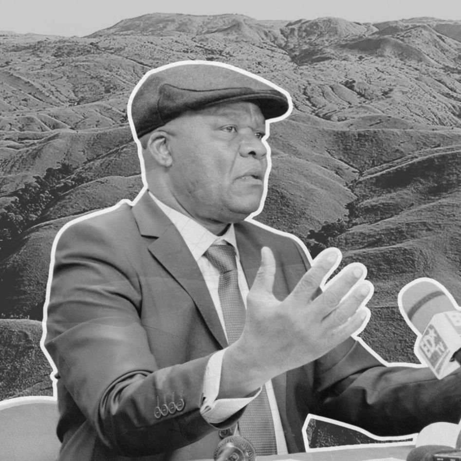 The image features a man in a suit and a cap, gesturing while speaking at a press conference or similar setting. He appears to be in front of a landscape of rolling hills or mountains, which are shown in a monochrome style to highlight the subject. The background scenery emphasizes the natural environment, contrasting with the formal attire of the man, who seems to be making an important point. The overall composition combines elements of nature and a formal discussion.