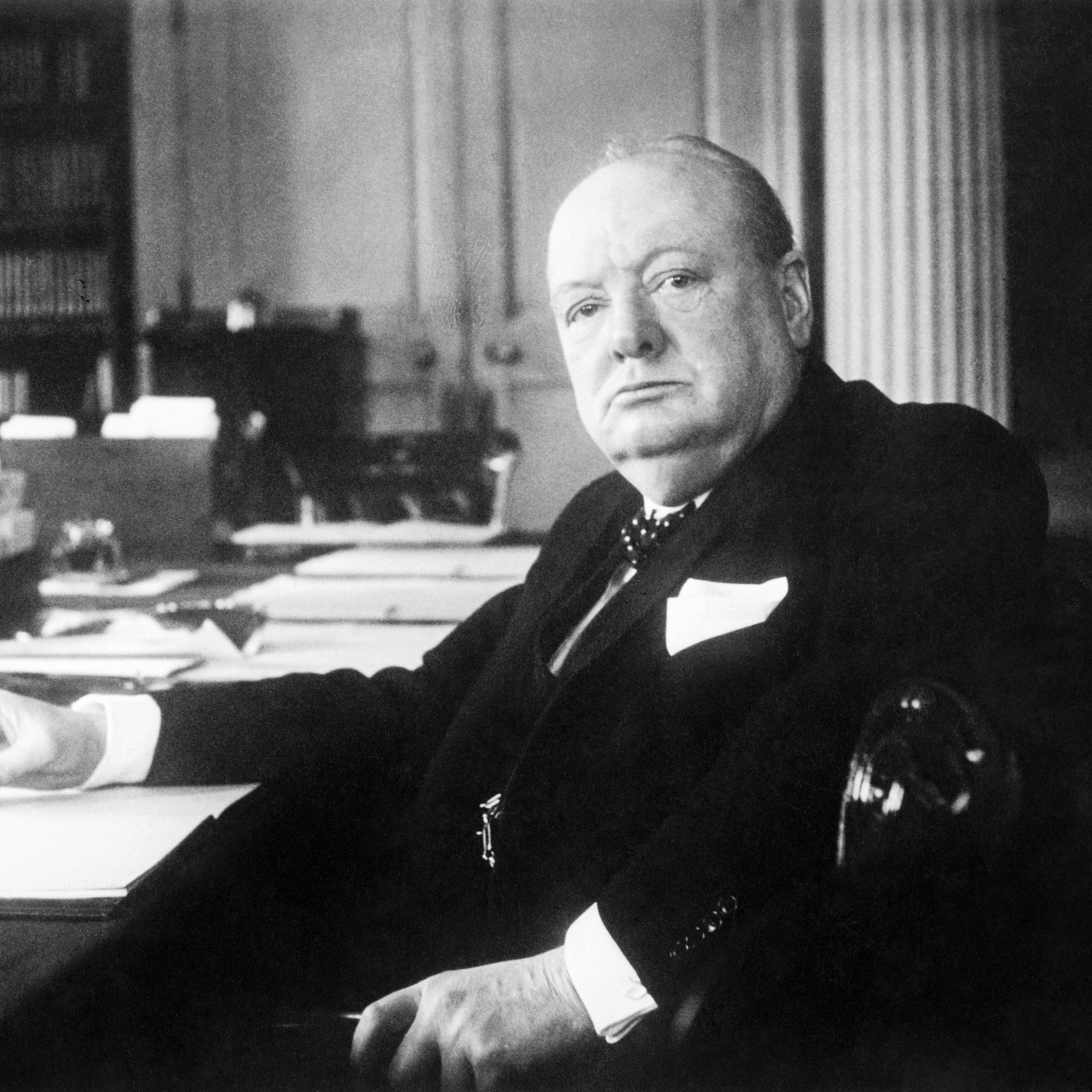 L'image présente un homme assis à un bureau. Il a une expression sérieuse et regarde droit devant lui. Son visage est rond et bien en chair, avec des traits marqués. Il est vêtu d'un costume sombre, avec une chemise claire et une cravate. Dans sa main, il tient un stylo, et devant lui, des feuilles de papier sont disposées sur le bureau. En arrière-plan, on distingue des étagères remplies de livres. L'atmosphère de l'image suggère une ambiance de travail studieuse et normalement formelle.