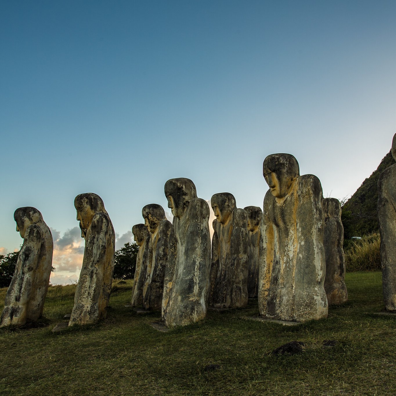 The image features a series of large stone sculptures arranged in a row, resembling human figures. These sculptures have distinct, elongated shapes and facial features, with some appearing to look downward. The background includes a grassy area and a hillside, with trees visible nearby. The sky above is clear, transitioning from blue to lighter tones as it approaches the horizon, suggesting either dawn or dusk. The overall atmosphere gives a sense of mystery and historical significance to the figures.