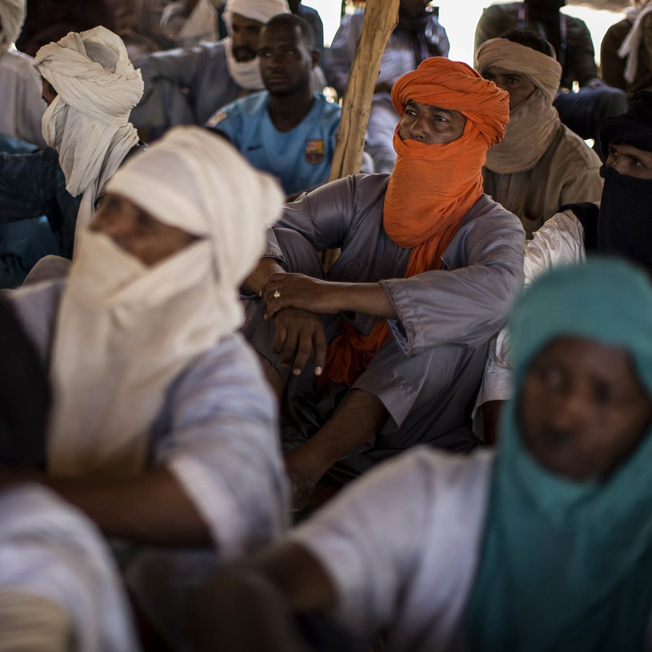L'image montre un groupe de personnes assises dans un espace clos. Les individus portent des vêtements traditionnels, souvent en tissu léger, et sont partiellement masqués par des turbans ou d'autres pièces de tissu. On peut percevoir des couleurs variées : certains portent des turbans orange, d'autres bleus ou verts. Les visages des personnes sont en grande partie cachés, créant une atmosphère de mystère. L'environnement semble calme, et les positions des corps indiquent une attention collective. La lumière est douce, suggérant un éclairage naturel venant d'une source extérieure. On peut également sentir une ambiance de rassemblement, possiblement pour un échange culturel ou une discussion importante.