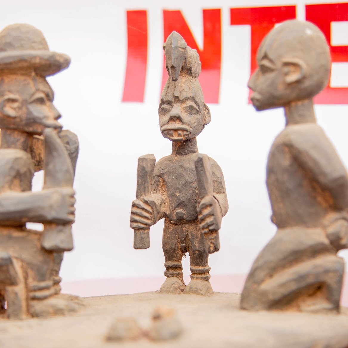 L'image présente une sculpture en bois représentant trois figures humaines stylisées. Au centre, une figure masculine, aux traits marqués, tient un objet dans les mains, probablement un outil ou un symbole culturel. Il est entouré de deux autres sculptures, l'une à gauche, qui porte un chapeau large, et l'autre à droite, avec une coiffure distinctive. Les personnages semblent engagés dans une conversation ou un échange, ce qui suggère une interaction sociale. Les détails des sculptures, comme les expressions des visages et les postures, témoignent d'un art tribal riche en histoire et en culture. Le fond de l'image est flou, mettant l'accent sur les sculptures elles-mêmes.