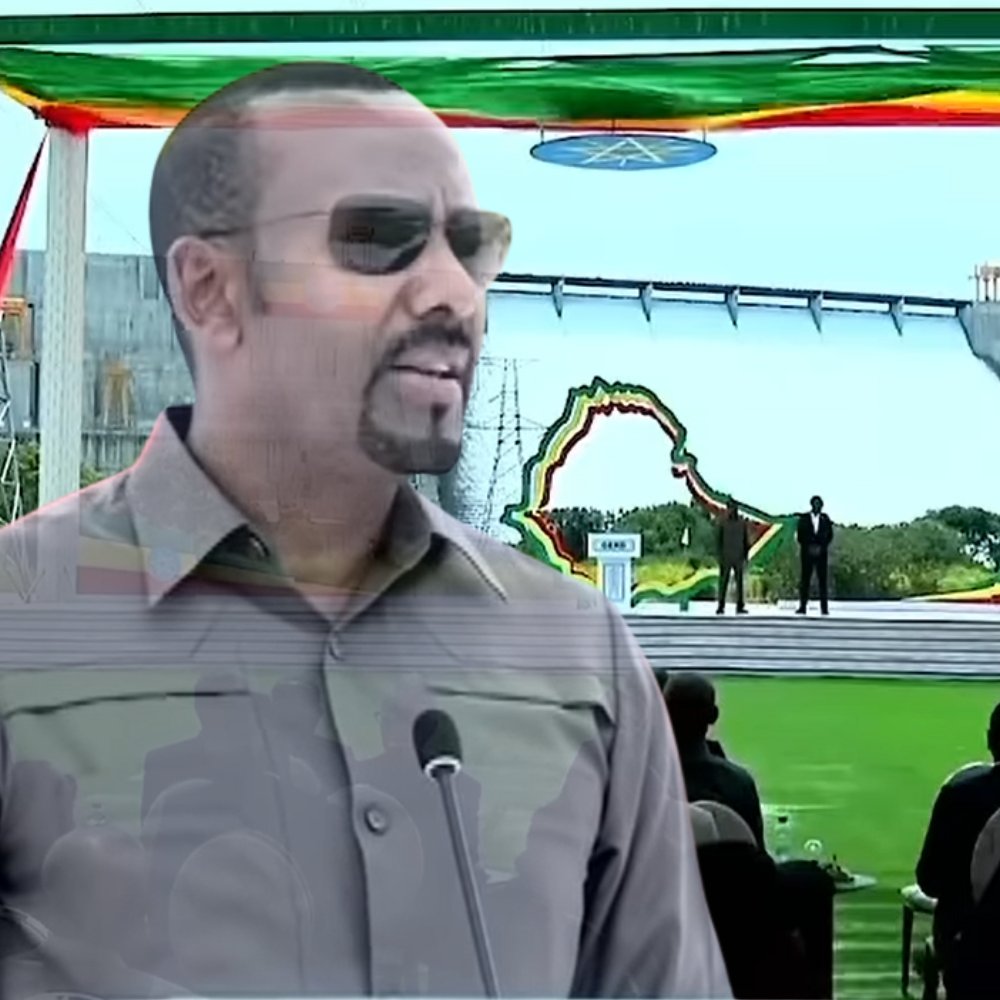 L'image montre un homme debout au premier plan, portant des lunettes de soleil et un uniforme militaire, qui semble s'adresser à un public. Il est probablement en train de faire un discours. En arrière-plan, on aperçoit une grande scène décorée de drapeaux éthiopiens et d'autres couleurs vives, avec une grande structure ressemblant à un barrage. Au premier plan, on peut voir des personnes assises, attentives, apprêtées pour un événement officiel en plein air, sur une pelouse verte. L'atmosphère semble d'une grande importance, peut-être liée à une célébration ou une inauguration.