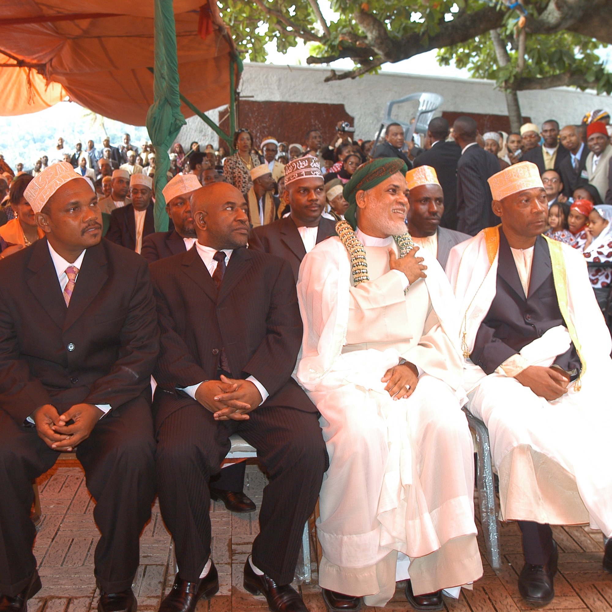 L'image montre un groupe d'hommes assis côte à côte lors d'un événement en plein air. Au premier plan, quatre hommes bien habillés portent des costumes sombres et des chapeaux traditionnels. Le premier homme à gauche a des lunettes et affiche une expression sérieuse. Le deuxième, au milieu, est habillé en noir et semble concentré. Le troisième, portant une robe traditionnelle blanche et un turban, sourit et semble être en conversation. Le quatrième homme, également en tenue traditionnelle, regarde droit devant lui. En arrière-plan, on peut apercevoir une foule de personnes rassemblées, avec des arbres et des tentes fournissant de l'ombre. L'ambiance paraît formelle et respectueuse, suggérant que ce moment est empreint de signification culturelle ou de cérémonie importante.