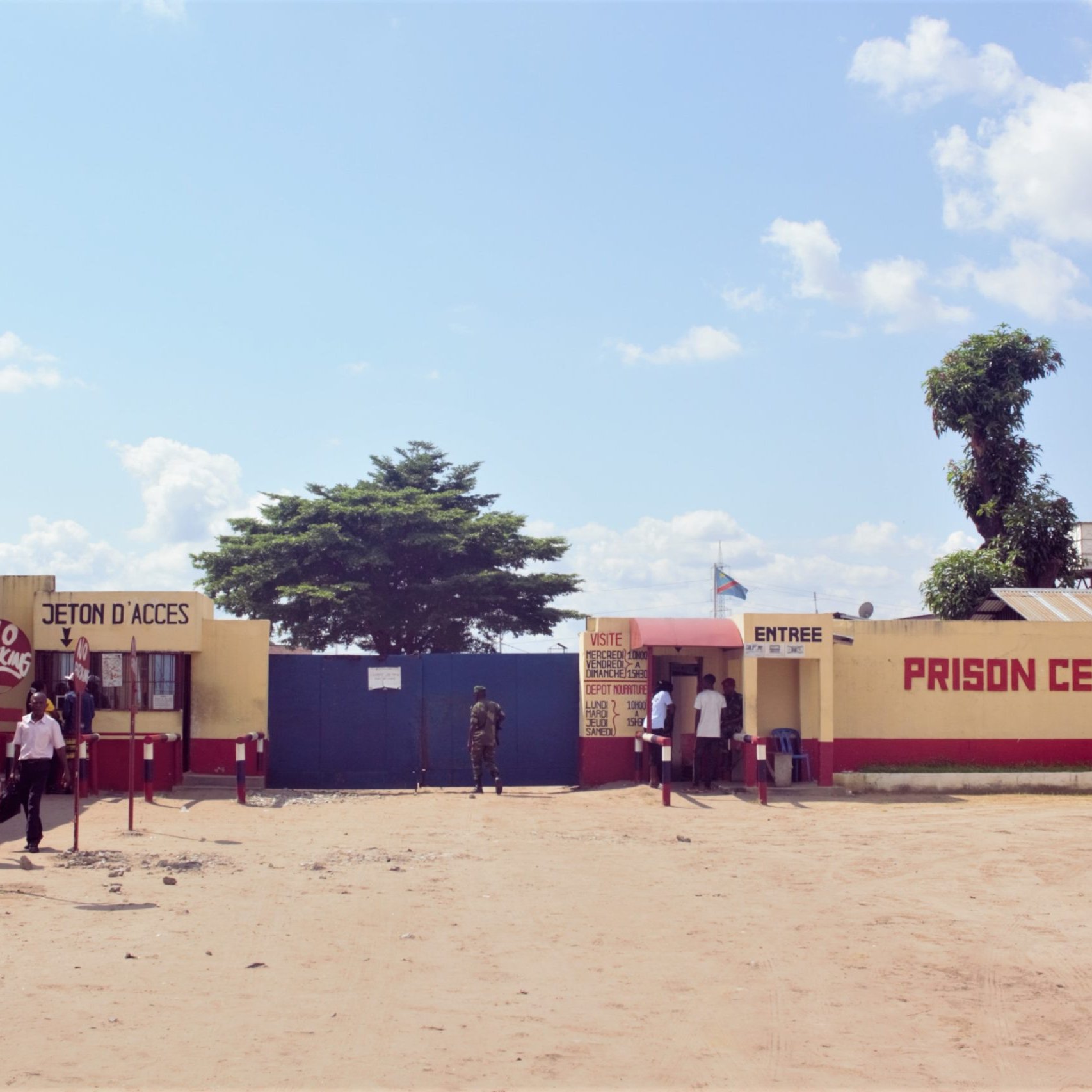 L'image montre l'entrée de la Prison Centrale de Makala. Devant le bâtiment, on voit une grande porte bleue. À droite, une cabine d'accès avec des petits guichets, probablement pour les visiteurs. Des personnes sont sur le point d'entrer ou de sortir. L'environnement est désertique, avec du sable au sol. Quelques voitures sont garées à droite, et un grand arbre se dresse à proximité. Le ciel est clair, avec quelques nuages. Des inscriptions sont visibles sur les murs, notamment le nom de la prison.