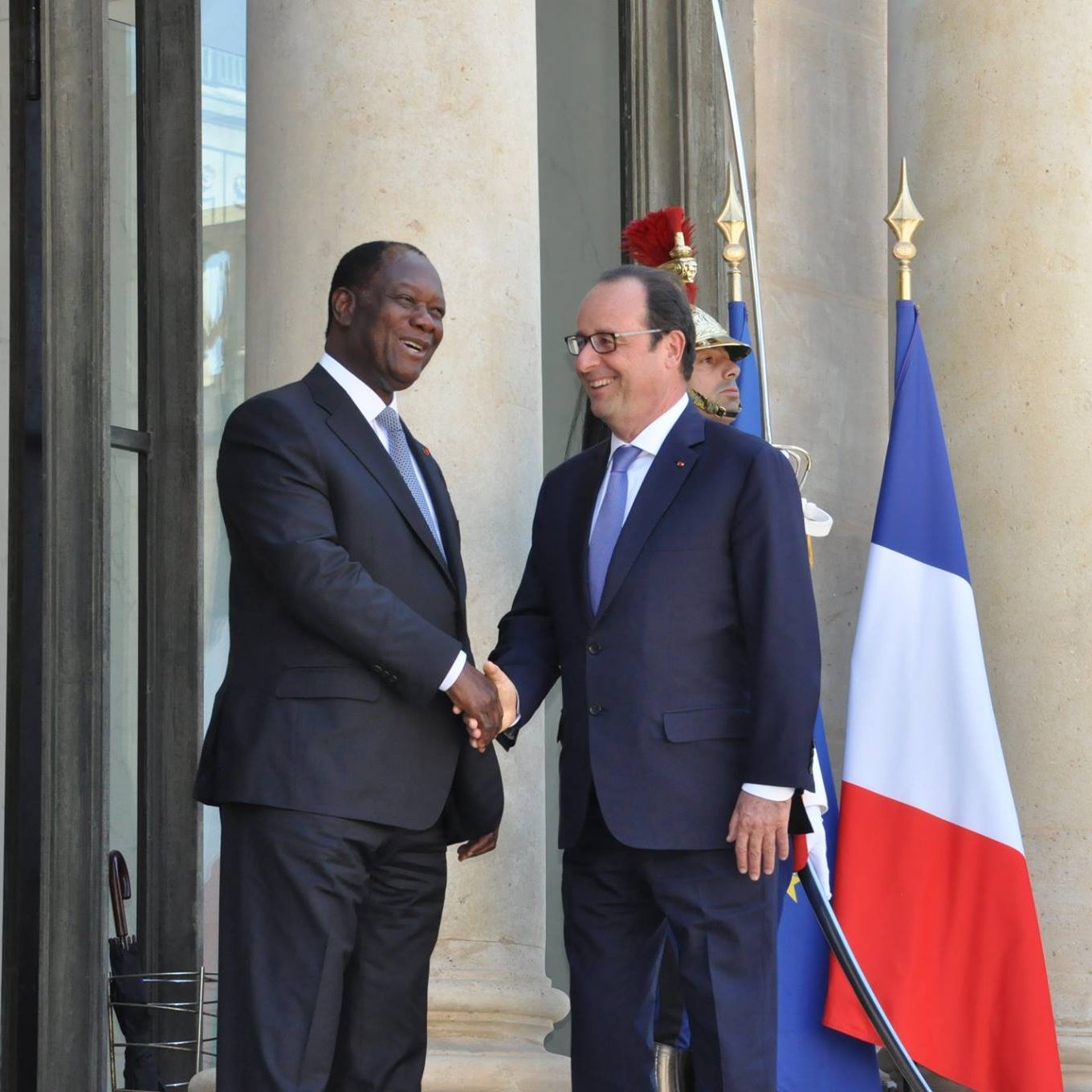 L'image montre deux hommes se serrant la main sur un perron, devant un bâtiment imposant. L'un d'eux porte un costume sombre avec une cravate, tandis que l'autre est vêtu d'un costume clair. En arrière-plan, on aperçoit un drapeau français tricolore et des gardes en uniforme militaire. Le décor suggère une atmosphère de rencontre officielle et de diplomatie. Les colonnes du bâtiment ajoutent une touche de majesté à la scène.