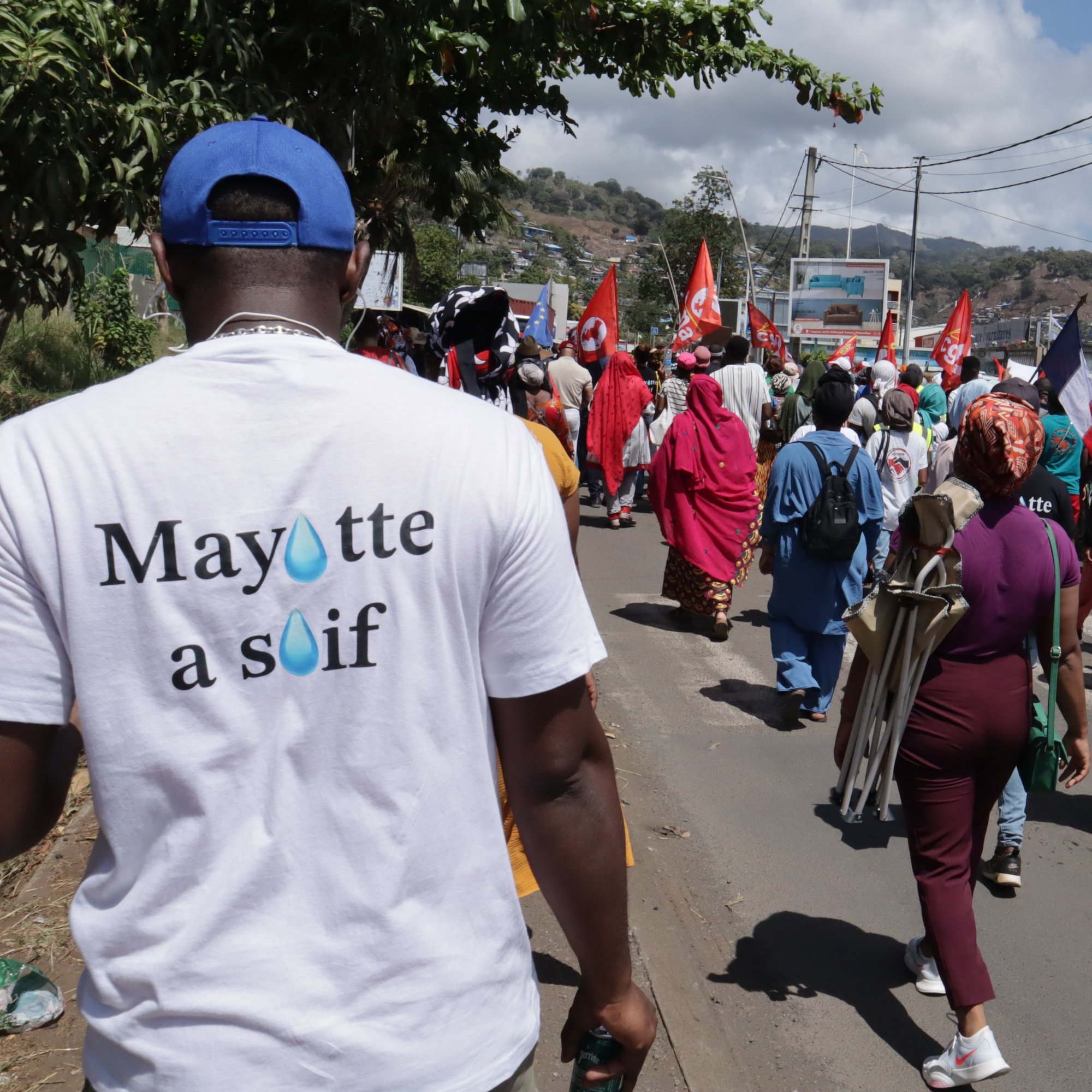 La scène dépeint un rassemblement animé dans une rue, où un groupe de manifestants avance. Au premier plan, on aperçoit une personne portant un t-shirt blanc avec l'inscription "Mayotte à souff" sur le dos. Les manifestants, vêtus de couleurs variées, portent des drapeaux rouges et d'autres pancartes, créant une atmosphère de solidarité et de revendication. À l'arrière-plan, on voit des bâtiments et des montagnes, sous un ciel partiellement nuageux. Les sons de la foule, les discussions et les chants de protestation résonnent, ajoutant à l'énergie de l'événement.