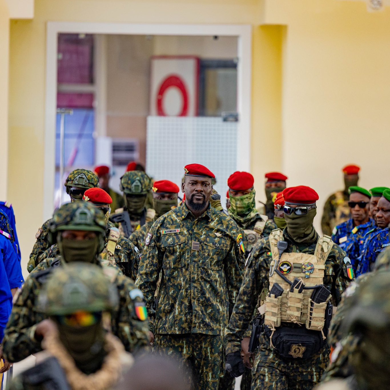 L'image représente un groupe de militaires rassemblés dans un intérieur lumineux. Au centre, un homme en uniforme se distingue par son attitude imposante. Il porte un camouflage militaire et un béret rouge. Les autres soldats autour de lui sont vêtus d'uniformes similaires, certains portant des masques de camouflage. Leurs bérets varient en couleur, notamment le rouge et le vert, créant un contraste visuel. L'ensemble évoque une atmosphère de discipline et de solidarité au sein des forces armées.