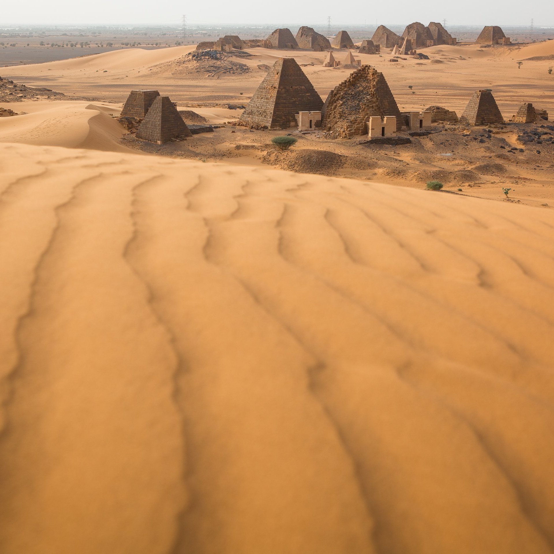 L'image décrit un paysage désertique, dominé par de vastes dunes de sable aux formes ondulantes. En arrière-plan, on aperçoit des pyramides anciennes dont les sommets sont pointus et les surfaces sont qui semblent rugueuses. Le terrain autour des pyramides est également parsemé de petites éboulis et de dunes, créant un paysage à la fois majestueux et mystérieux. La lumière du soleil éclaire la scène, mettant en valeur les nuances dorées du sable et l'architecture historique des pyramides. L'atmosphère générale évoque la chaleur et l'immensité du désert, ainsi qu'un sentiment d'émerveillement face aux vestiges du passé.