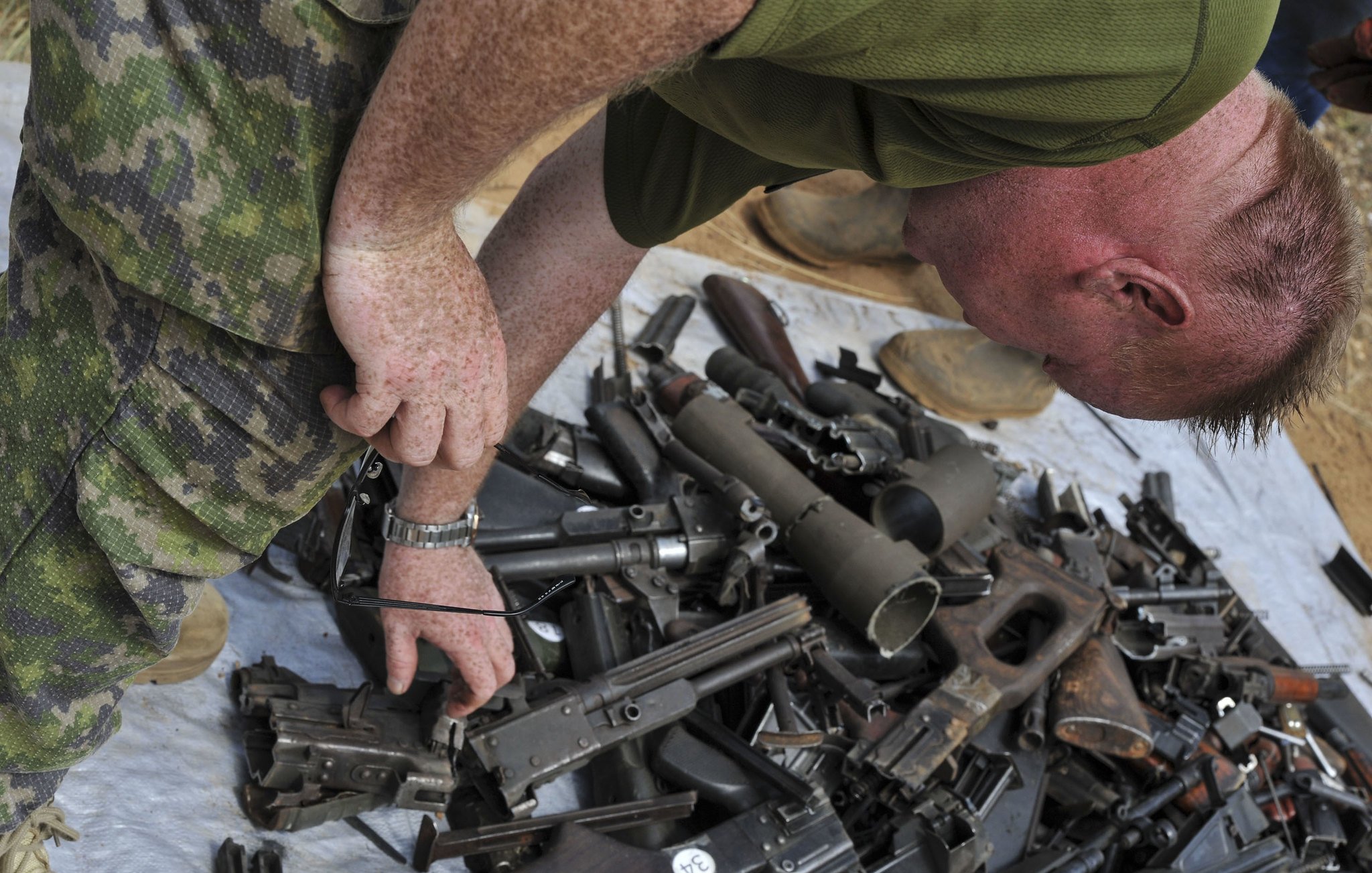 Dans cette image, on peut voir un homme en tenue militaire, penché en avant, qui examine un tas d'armes. Ce tas, étalé sur une surface comme une bâche ou un sol dur, est composé de diverses armes à feu, incluant des fusils et des pistolets, qui sont éparpillés de manière désordonnée. L'homme, avec des cheveux courts et roux, semble concentré dans sa tâche d'inspection. L'environnement autour de lui pourrait évoquer un contexte militaire ou de désarmement.