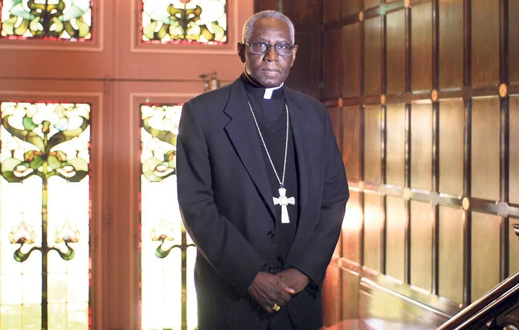 Dans cette image, nous voyons un homme debout dans un environnement qui semble être une pièce élégante, probablement une église ou un bureau religieux. Il porte une tenue noire, typique des membres du clergé, avec une soutane et un col ecclésiastique. Autour de son cou, une grande croix pend, symbolisant son rôle religieux. Le fond est composé de murs en bois foncé et de vitraux colorés, ce qui crée une atmosphère solennelle et respectueuse. Les motifs des vitraux laissent transparaître une lumière douce, ajoutant une dimension spirituelle à l'ensemble de la scène. L'homme a une expression sérieuse, se tenant droit et digne, ce qui évoque l'autorité et la sagesse.
