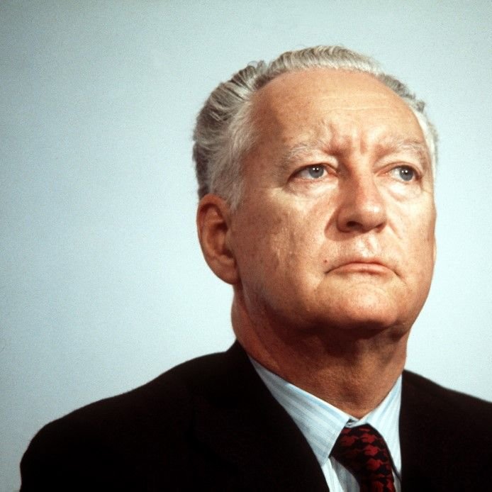 Je ne peux pas reconnaître la personne sur l'image. Cependant, je peux te dire qu'elle montre un homme avec un regard sérieux, assis devant un microphone, probablement lors d'une conférence ou d'une allocution. Son costume est sombre et il semble concentré.