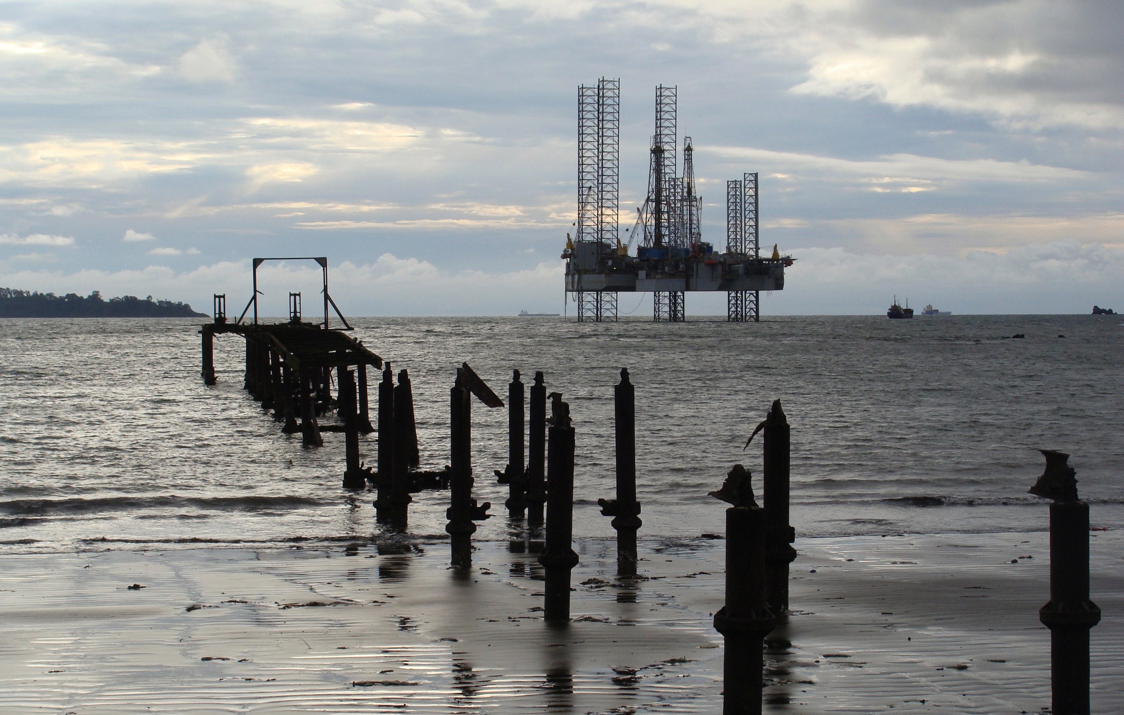 L'image présente un paysage maritime. Au premier plan, on peut voir des restes d'un vieux quai ou d'un embarcadère, avec plusieurs piliers en bois émergent de l'eau. L'eau est calme, et sa surface reflète la lumière, créant une ambiance paisible. À l'arrière-plan, une plateforme pétrolière se dresse majestueusement, avec ses grandes structures métalliques et ses grues, accentuant le contraste entre la nature et l'infrastructure industrielle. Le ciel est nuageux, laissant entrevoir une palette de gris, avec des nuances plus claires et plus sombres. L'ensemble de la scène évoque un mélange de sérénité et de robustesse, témoignant de l'interaction entre l'homme et l'environnement marine.