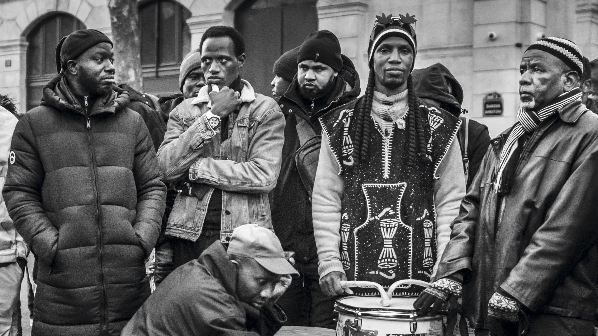 L'image montre un groupe de plusieurs hommes rassemblés dans un environnement urbain. Ils portent des vêtements variés, allant de parkas sombres à des vêtements traditionnels. Certains d'entre eux regardent dans des directions différentes, affichant une diversité d'expressions. Au centre, un homme est assis, jouant sur un tambour, ajoutant une ambiance musicale à la scène. Les autres semblent attentifs, créant une atmosphère de camaraderie et d'échange. L'image est en noir et blanc, accentuant les contrastes et les textures des vêtements et des visages.