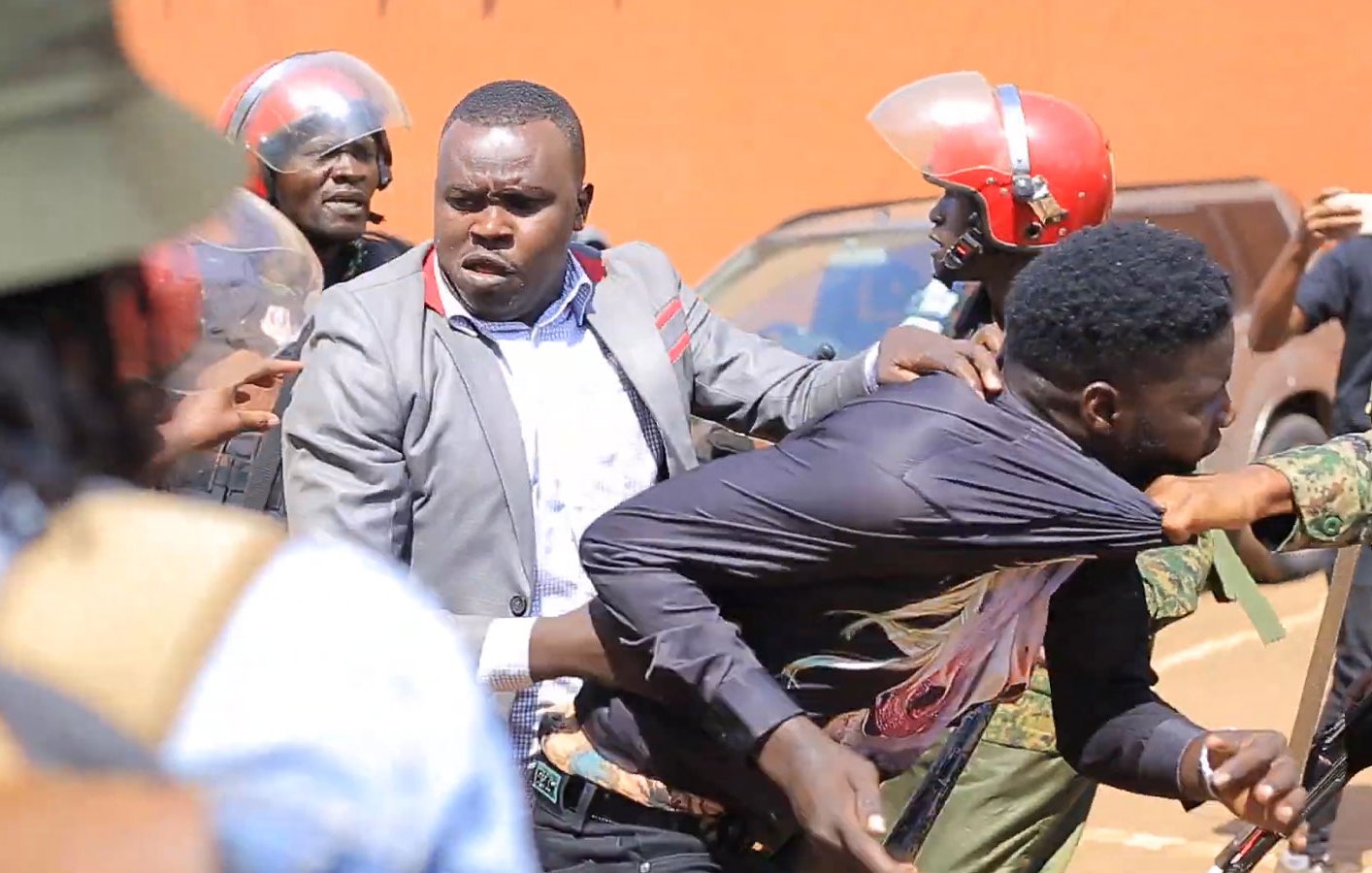 L'image montre une scène tendue sur une rue. On peut voir des policiers en uniforme, portant des casques rouges, qui semblent intervenir auprès d'un homme. Ce dernier est tiré en arrière par un autre homme qui essaie de le protéger. L'atmosphère est chaotique, avec des tensions visibles entre les forces de l'ordre et la personne qu'ils maîtrisent. En arrière-plan, il y a quelques personnes observant la situation et des véhicules garés. Le paysage évoque une ambiance de manifestation ou d'émeute.