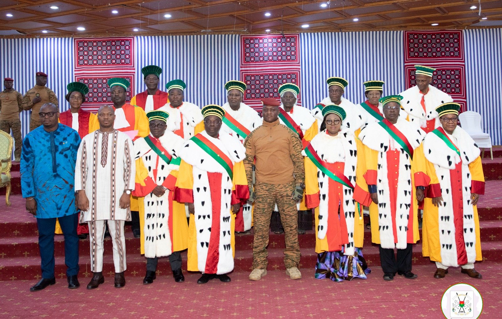 The image features a formal gathering of a group of individuals, likely in a ceremonial setting. They are wearing traditional attire, which includes colorful robes adorned with striped and patterned elements. Many of them are also wearing distinctive hats or headgear. The setting appears to be well-decorated, possibly for an official event, with a red carpet and banners or decorations in the background. A few individuals stand out in military or official uniforms, suggesting a mix of civilian and military presence at the event.