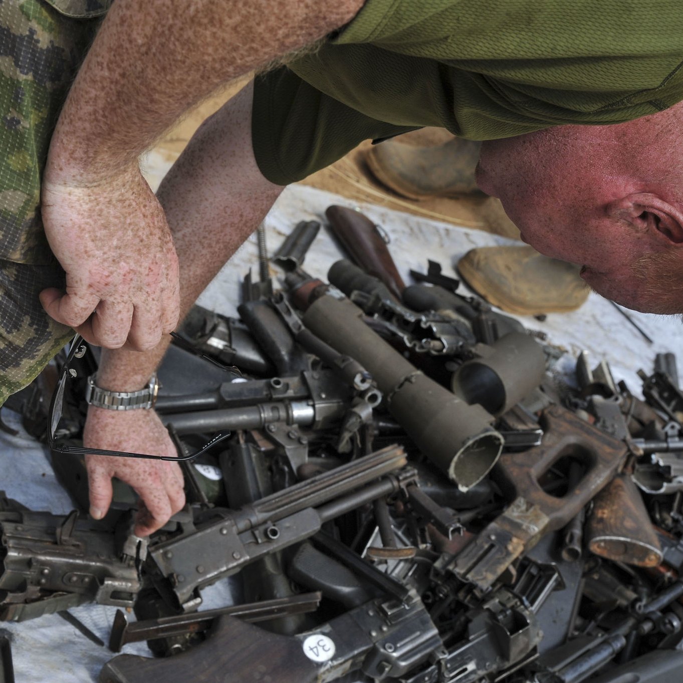 Dans cette image, on peut voir un homme en tenue militaire, penché en avant, qui examine un tas d'armes. Ce tas, étalé sur une surface comme une bâche ou un sol dur, est composé de diverses armes à feu, incluant des fusils et des pistolets, qui sont éparpillés de manière désordonnée. L'homme, avec des cheveux courts et roux, semble concentré dans sa tâche d'inspection. L'environnement autour de lui pourrait évoquer un contexte militaire ou de désarmement.