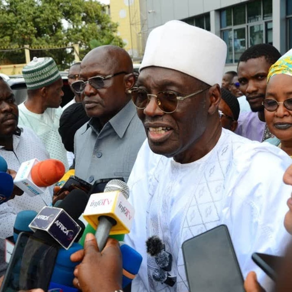 Dans cette image, un groupe de personnes se trouve en train de discuter avec des journalistes. Au centre, un homme porte un costume traditionnel blanc, accompagné d'un chapeau traditionnel et d'une écharpe. Il parle aux médias, entouré de plusieurs personnes, dont certaines semblent prendre des notes ou poser des questions avec des micros devant eux. Les visages des personnes montrent un mélange d'intérêt et de concentration. En arrière-plan, on peut apercevoir un bâtiment moderne et des arbres, ce qui donne une ambiance urbaine à la scène.