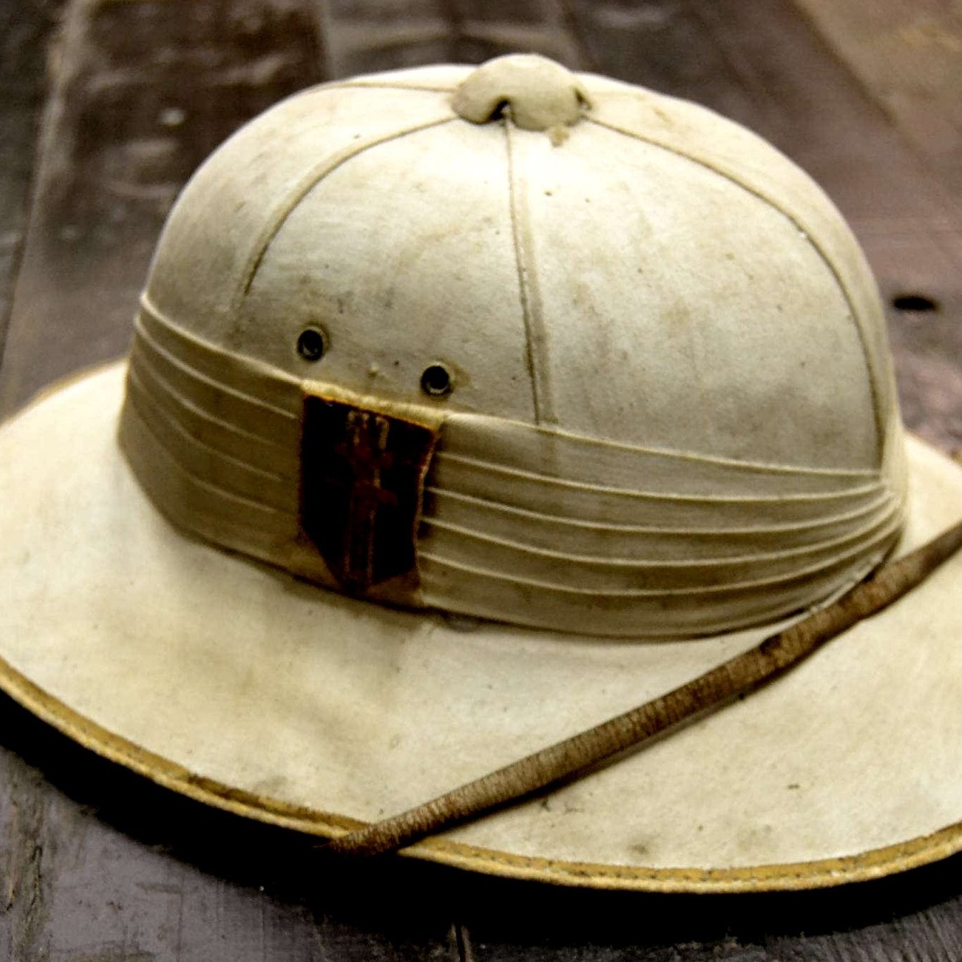 L'image montre un chapeau de style explorer, souvent associé aux expéditions dans des régions chaudes ou tropicales. Le chapeau est de couleur claire, presque beige, avec une large bordure qui protège du soleil. Sa partie supérieure est légèrement surélevée, indiquant une forme arrondie. À l'avant, il y a une bande de tissu plissé avec un morceau de bois ou de cuir, donnant un aspect rustique. Une fine tige est posée sur le chapeau, ajoutant une touche naturelle. La surface du chapeau semble légèrement usée, témoignant d'un usage antérieur. Le fond sur lequel il repose est en bois, ce qui renforce l'atmosphère d'aventure.