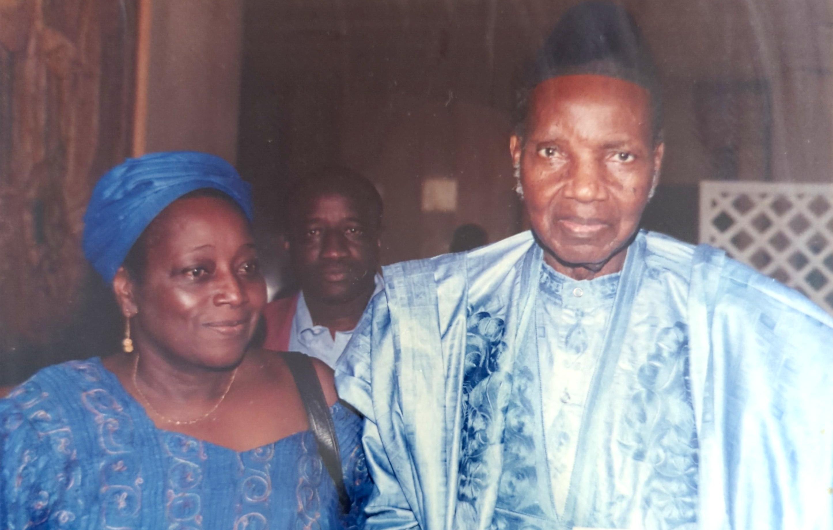 L'image montre trois personnes. À l'avant, un homme et une femme sont vêtus de costumes traditionnels, probablement en tissus d'importance culturelle. L'homme porte un chapeau et a un regard sérieux, tandis que la femme a un sourire et semble détendue. En arrière-plan, un troisième homme est visible, bien qu'il soit moins en avant-plan. L'ambiance de l'image semble formelle.