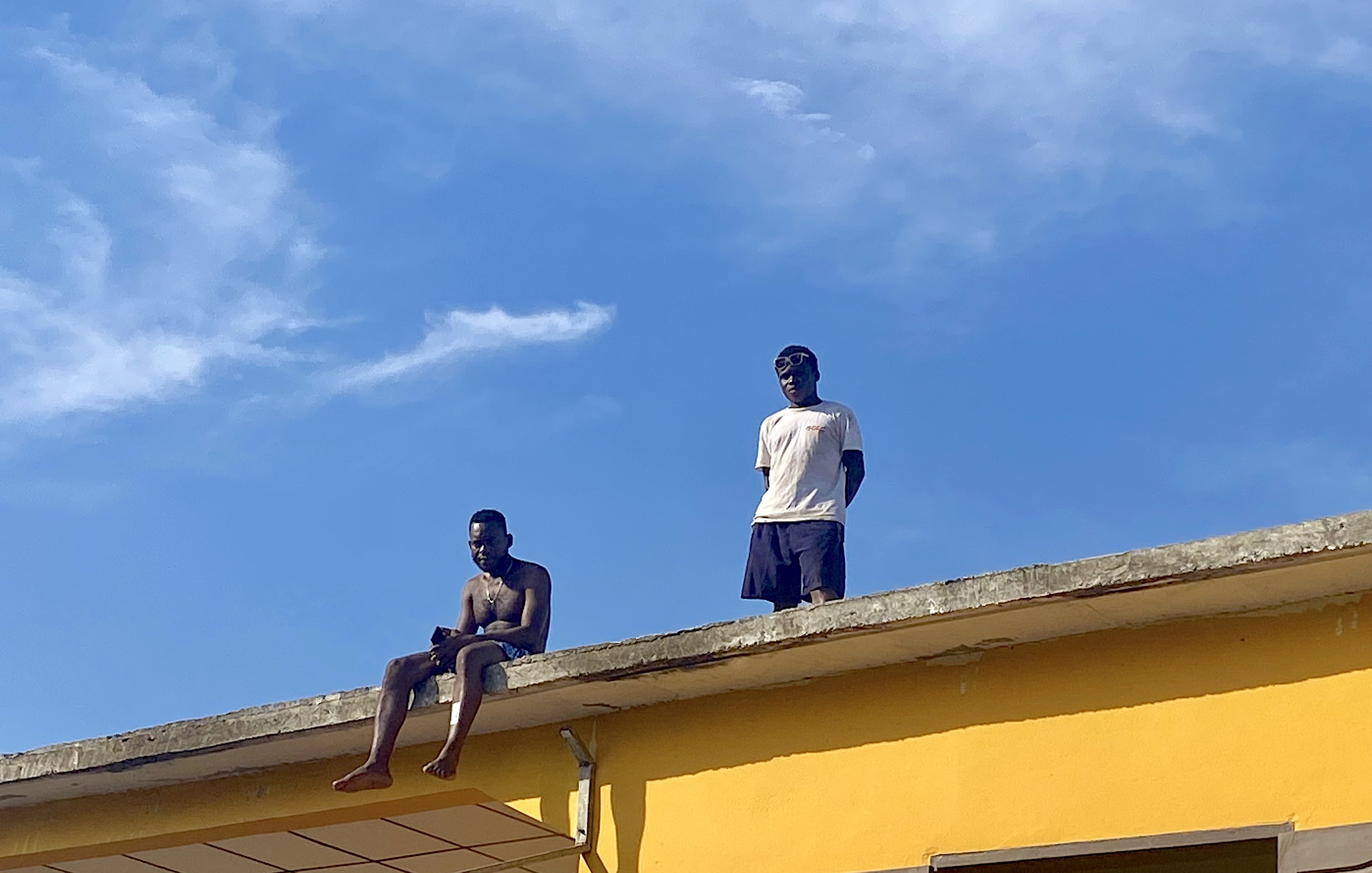 Sur cette image, deux jeunes hommes se trouvent sur le toit d'une maison peinte en jaune vif. L'un d'eux est assis au bord, les jambes pendantes dans le vide, tandis que l'autre se tient debout à ses côtés. Le ciel est clair et bleu, avec quelques nuages légers. L'ambiance semble détendue, peut-être un moment de loisirs ou de camaraderie. Les détails de l'environnement autour d'eux ne sont pas visibles dans cette capture, mais l'accent est mis sur leur posture et l'interaction entre eux.