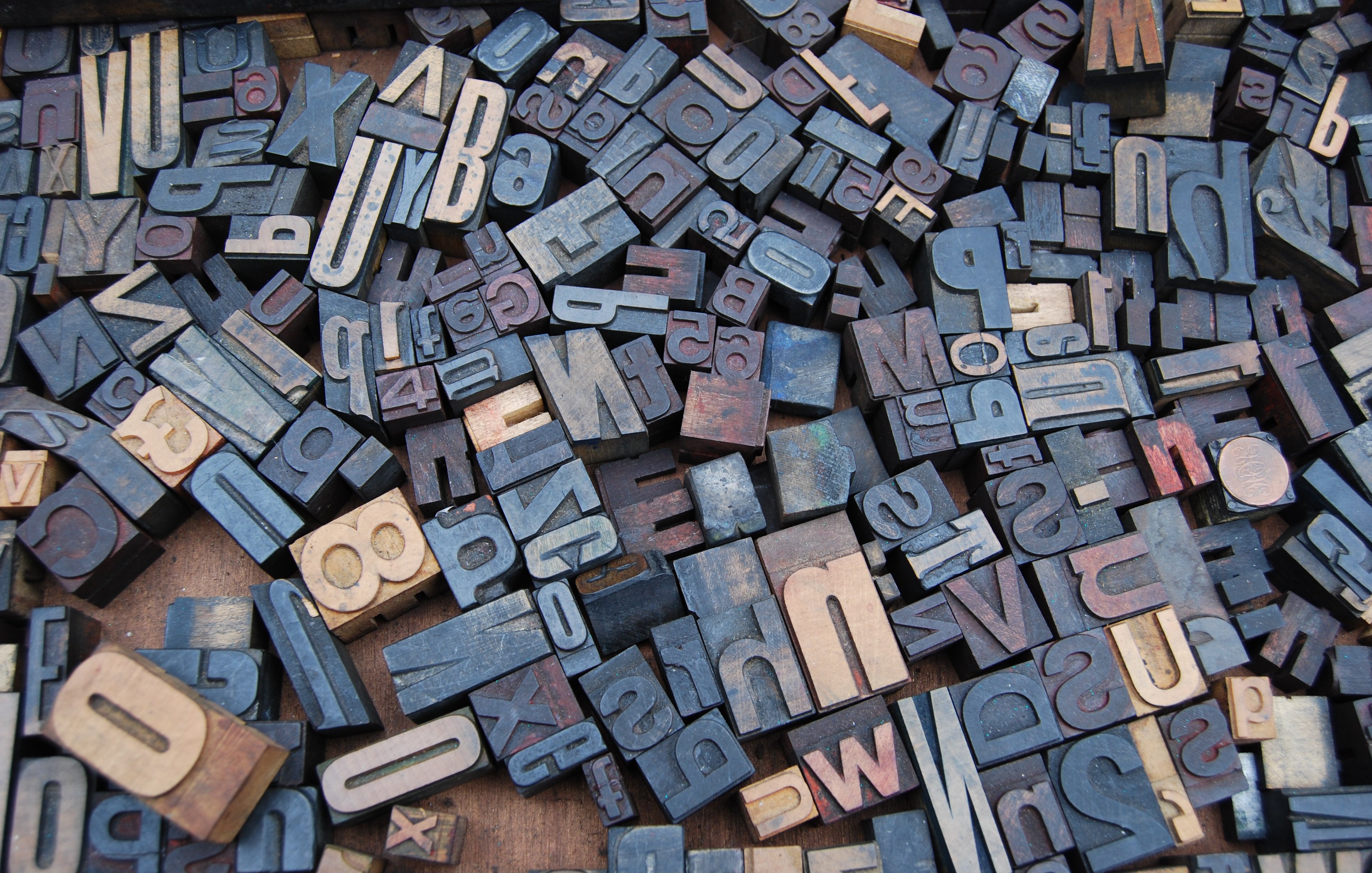 L'image montre une vaste collection de caractères typographiques en bois, disposés de manière apparemment aléatoire. Vous pouvez imaginer des lettres majuscules et minuscules, ainsi que des chiffres, chacun avec des motifs et textures variés. Certains caractères sont plus sombres, tandis que d'autres présentent des nuances plus claires, créant un contraste visuel intéressant. L'ensemble évoque une ambiance artisanale et rétro, rappelant l'époque de l'impression traditionnelle. L'atmosphère générale est celle d'un espace créatif, rempli de possibilités typographiques.