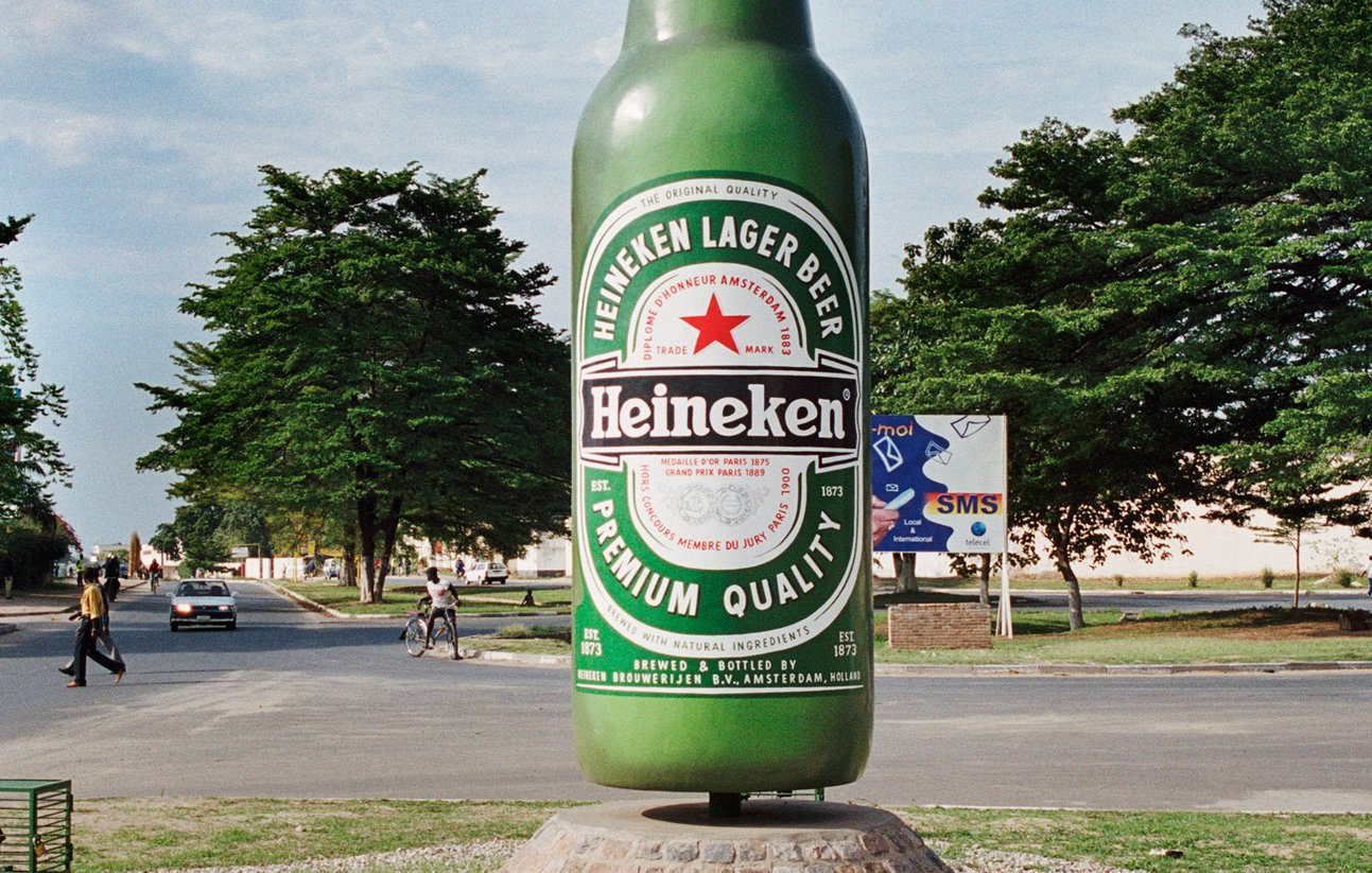 L'image montre une grande sculpture de bouteille de bière Heineken, qui se dresse au centre d'une intersection. La bouteille est peinte en vert vif et arborant l'étiquette rouge et blanche emblématique de la marque. Autour de la sculpture, on peut voir des gens se déplacer, certains à pied et d'autres à vélo. En arrière-plan, on distingue des arbres et quelques bâtiments qui ajoutent une ambiance locale. L'ensemble dégage une atmosphère animée et colorée, typique de l'urbanisme d'une ville vivante.