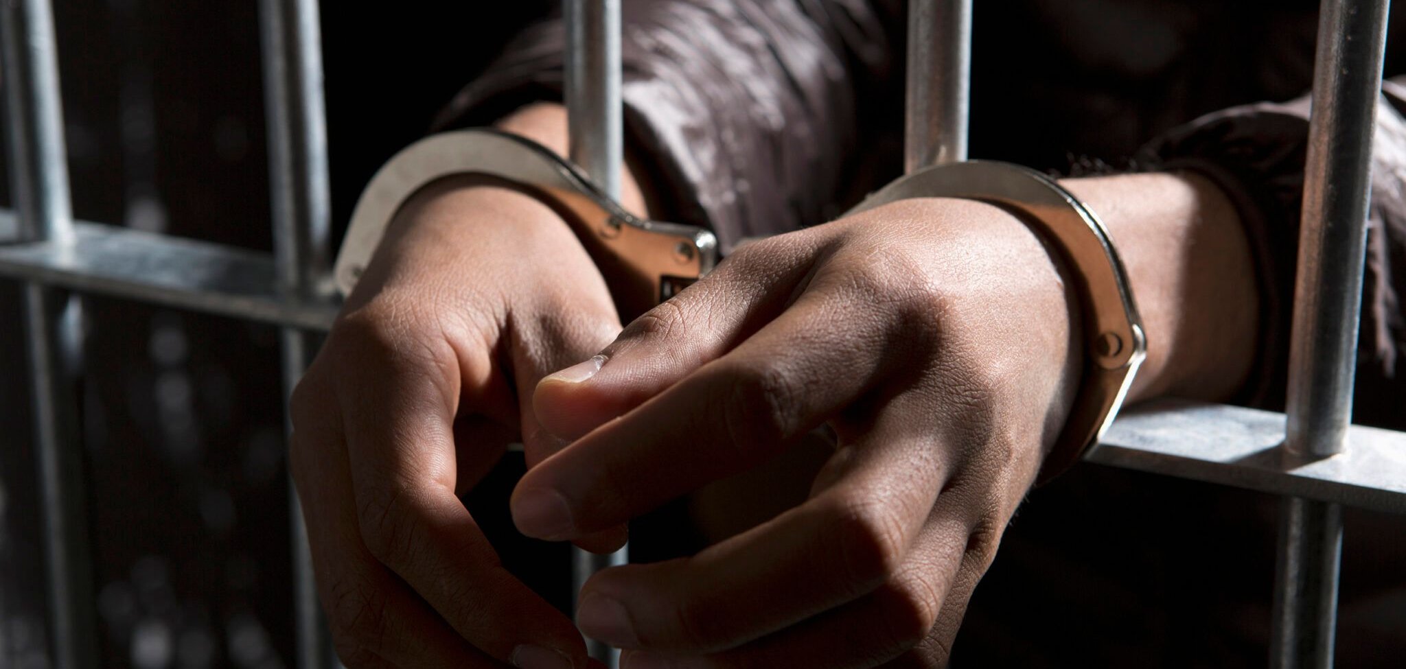 L'image montre des mains menottées, immobilisées par des menottes métalliques, qui se trouvent derrière des barreaux. Les mains sont tendues et légèrement fléchies, occupant une position qui évoque à la fois la vulnérabilité et la contrainte. Le fond est sombre, créant un contraste avec la lumière qui met en évidence les éléments métalliques des menottes et les détails de la peau. L'ensemble de la scène transmet une ambiance de confinement et de détention.