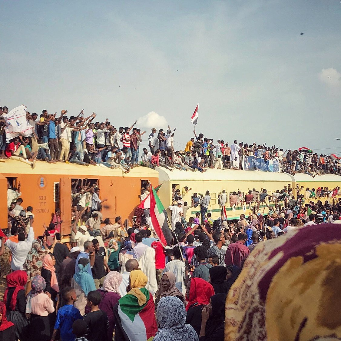 L'image montre une scène de grande effervescence, probablement lors d'un rassemblement ou d'une célébration. On peut voir une foule nombreuse, avec des gens debout sur un train, brandissant des drapeaux aux couleurs vives, en particulier du rouge, du noir et du vert. Les participants expriment leur enthousiasme et leur solidarité, certains levant les poings ou affichant des panneaux. La foule est diverse, incluant des personnes de tous âges, et il règne une atmosphère de ferveur collective, avec un ciel bleu parsemé de quelques nuages en arrière-plan. Cette image évoque des sentiments de joie, d'unité et de détermination.
