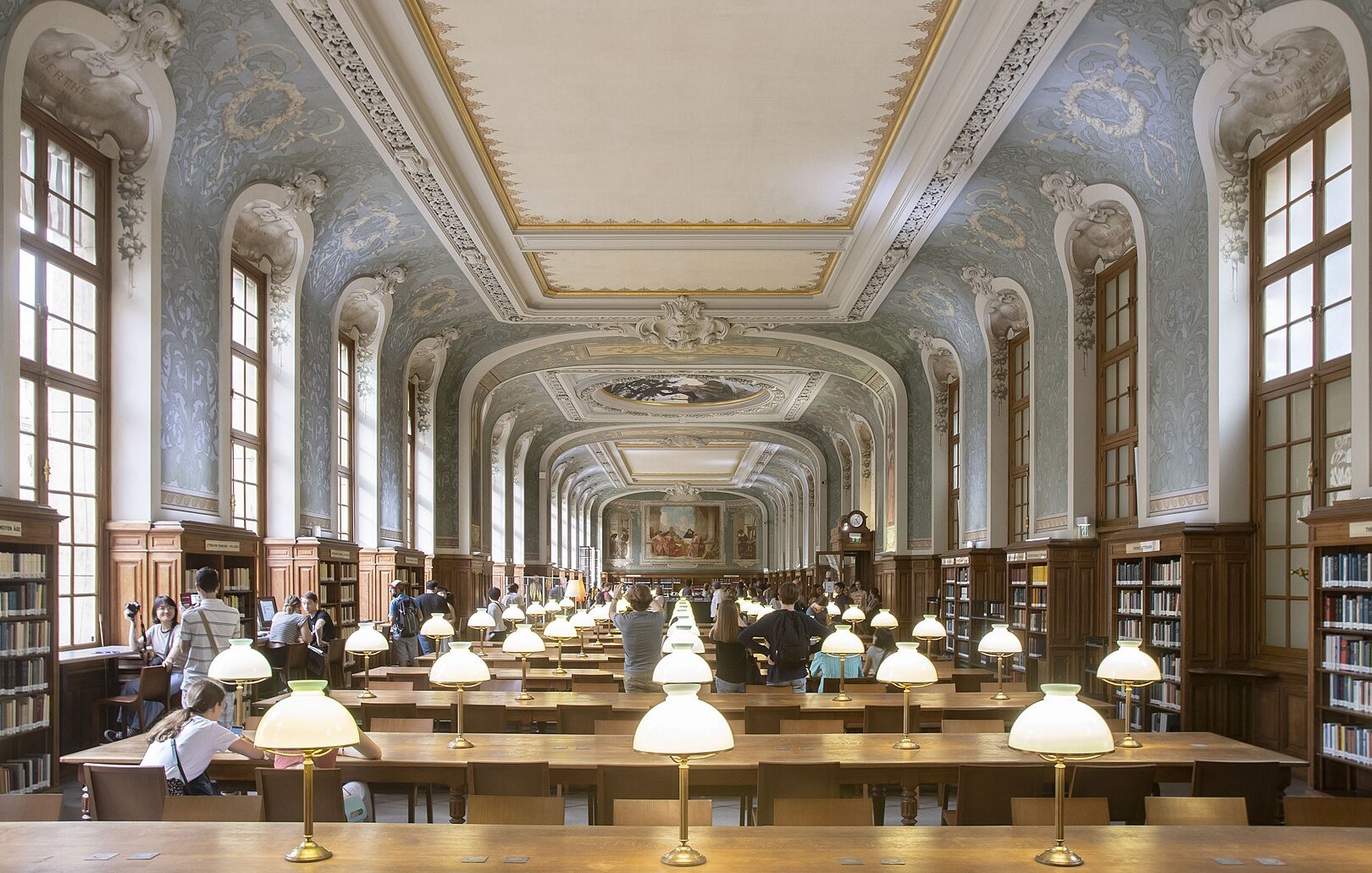 Imaginez une vaste bibliothèque, où l'air est chargé de silence et de connaissance. L'espace est long et majestueux, avec un plafond haut orné de décorations raffinées et de motifs artistiques. Les murs sont bordés de grandes fenêtres, permettant à la lumière naturelle d'inonder la pièce. Des lampes de table éclatent par leur lumière douce sur de longues tables en bois, disposées en rangées. Ces tables accueillent des visiteurs plongés dans leurs livres et leurs pensées. Au fond, un grand mur est décoré de fresques, ajoutant une note d'élégance à l'ensemble. Les étagères en bois, pleines de livres soigneusement rangés, s'élèvent majestueusement sur les côtés, évoquant un immense trésor de savoir à portée de main. Les gens se déplacent doucement, parlant à voix basse, ajoutant une ambiance de respect et de tranquillité. Cette bibliothèque semble être un sanctuaire pour les amateurs de lecture et de culture.