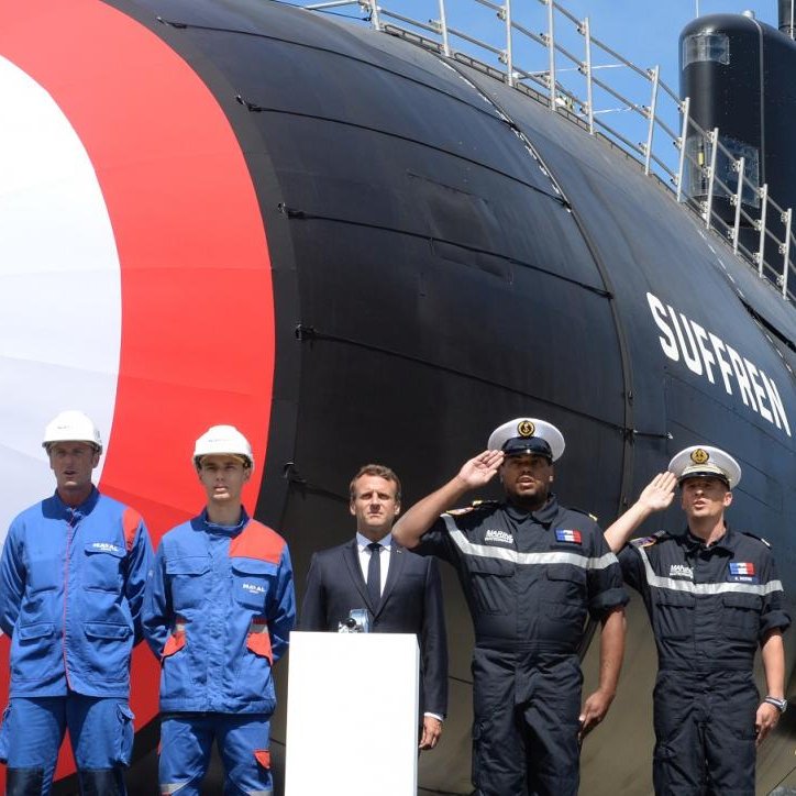 L'image montre un sous-marin de couleur noire avec un grand cercle rouge et bleu sur son côté. Devant le sous-marin, il y a quatre personnes en tenue de travail, dont deux en bleu et deux en uniforme noir. Elles se tiennent debout, affichant des expressions sérieuses. En arrière-plan, le ciel est clair et bleu, donnant une ambiance ensoleillée à la scène. Au centre, une plate-forme sur laquelle un homme en costume parle, probablement lors d'une cérémonie liée au sous-marin. Cela évoque une atmosphère de célébration ou d'inauguration d'un nouveau bateau.