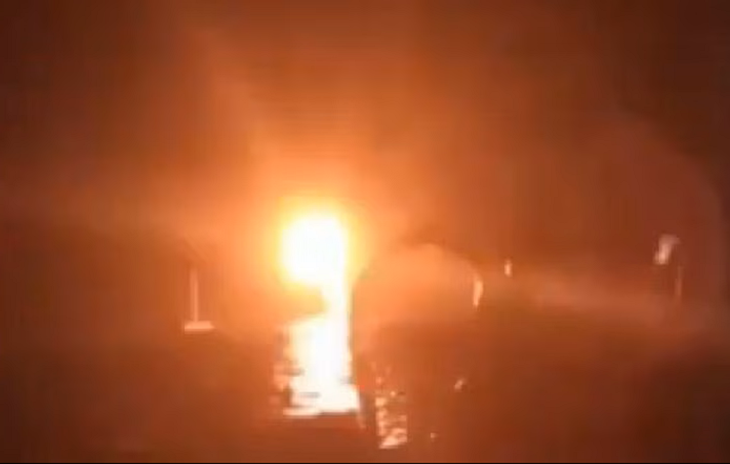 L'image présente une scène sombre avec une source de lumière éclatante au centre, semblant provenir d'un feu ou d'un soleil couchant. Les contours des objets et des formes sont flous, créant une ambiance mystérieuse. L'environnement pourrait évoquer un paysage naturel ou urbain, sans détails précis visibles. On ressent une intensité et une chaleur émises par cette lumière, contrastant avec l'obscurité environnante.