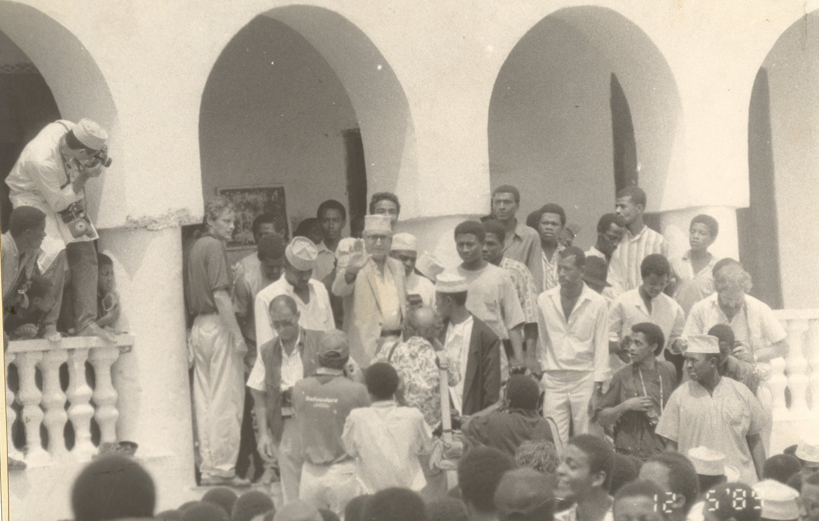 L'image représente une scène de rassemblement en extérieur, probablement lors d'un événement communautaire ou historique. Au premier plan, un groupe de personnes se regroupe autour d'une figure centrale, tandis qu'une autre personne, en haut à gauche, semble prendre une photo. Les participants portent des vêtements variés, certains avec des chapeaux. L'architecture en arrière-plan témoigne d'un style colonial, avec des arches et des balustrades. L'ambiance générale paraît vivante et joyeuse, suggérant un moment d'unité et de partage au sein d'une communauté.