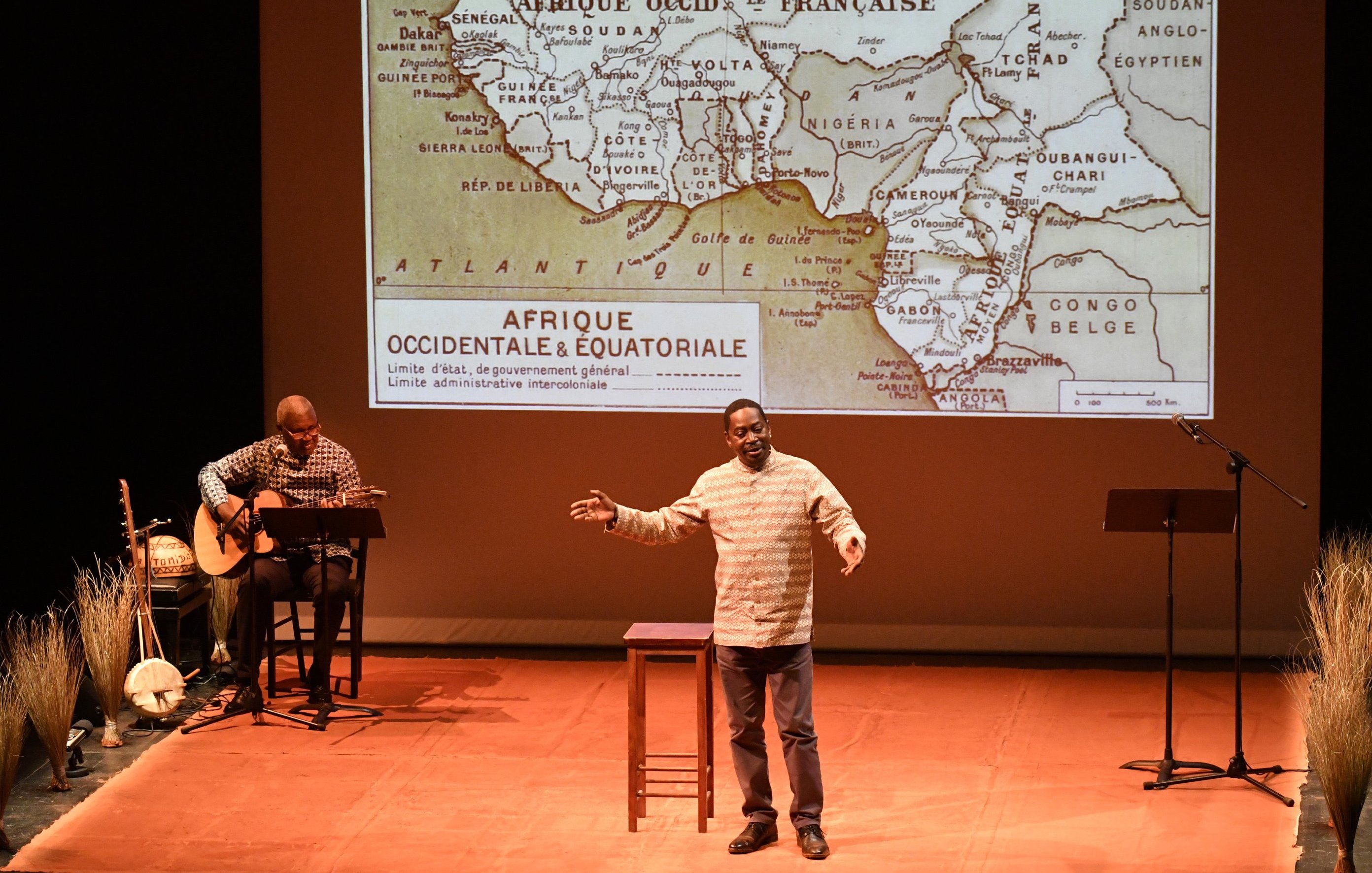 L'image représente un spectacle sur scène. Au premier plan, un homme s'exprime avec passion, les bras légèrement écartés pour accentuer son discours. Il porte une chemise claire et semble captivé par son sujet. À sa droite, un autre homme joue de la guitare assis sur un tabouret, ajoutant une ambiance musicale à la performance. En arrière-plan, un grand écran projette une carte ancienne de l'Afrique, spécifiquement d'Afrique occidentale et équatoriale, avec des contours de pays et des indications historiques. Le sol de la scène est décoré de brins de paille, ajoutant une touche naturelle à la mise en scène. L'éclairage doux met en valeur les acteurs et crée une atmosphère intimiste.