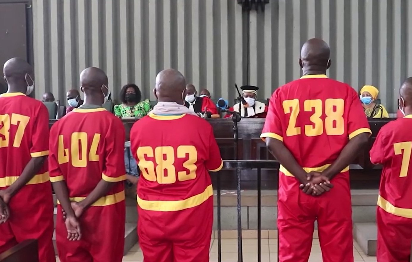 L'image montre un groupe de personnes dans un tribunal. Quatre hommes, portant des uniformes rouges numérotés au dos (337, 404, 683, 238, et 72), se tiennent debout, de dos, face à un juge et à un panel de personnes assises en face d'eux. L'atmosphère est sérieuse, et tous les participants portent des masques. Les murs du tribunal sont rayés, et on peut voir des membres de l'auditoire, y compris des femmes en vêtements colorés. L'ambiance générale évoque une situation judiciaire formelle.