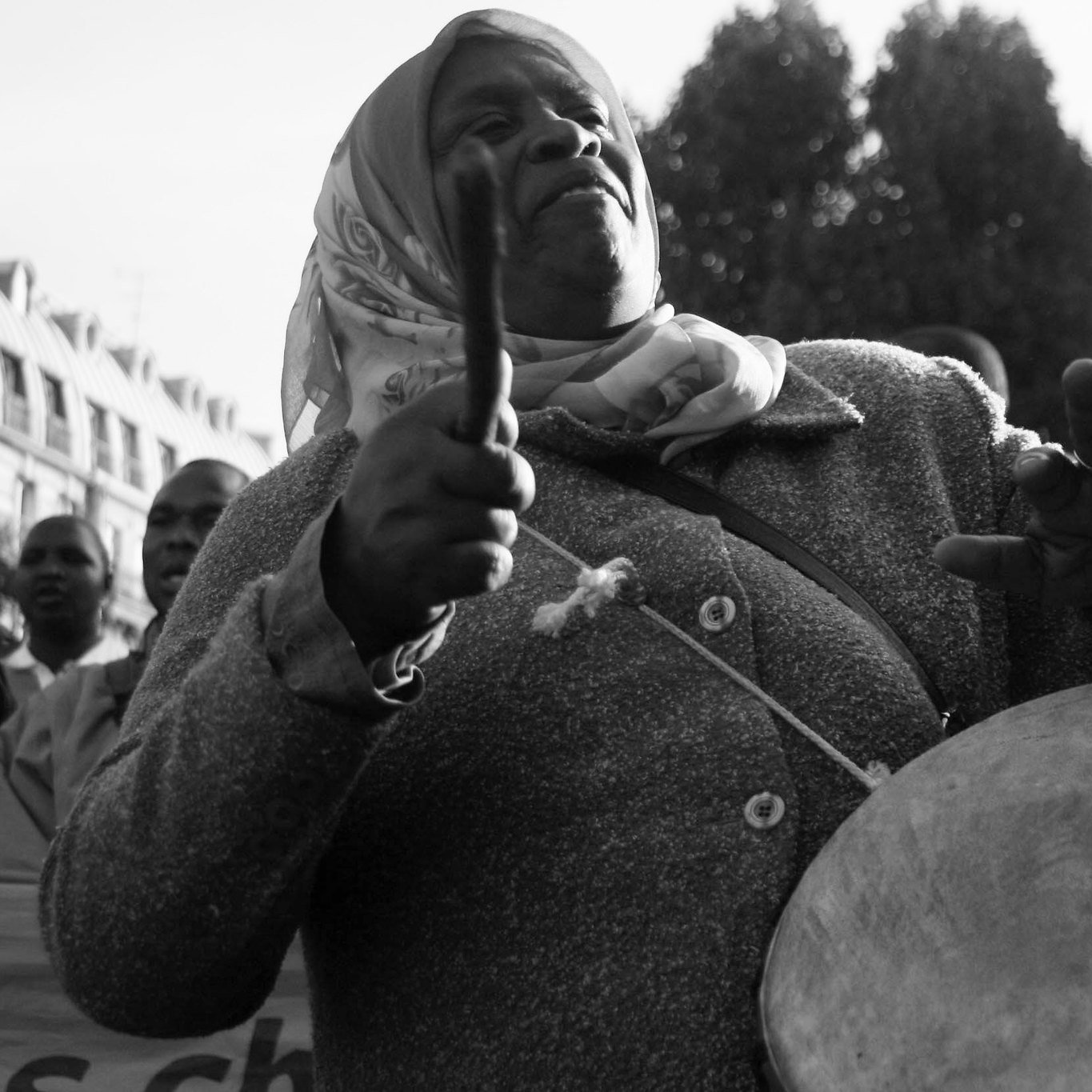 L'image représente une femme engagée dans un moment de mobilisation. Elle porte un manteau gris et un foulard autour de la tête. Ses bras sont levés, tenant des baguettes, et elle semble jouer d'un instrument de percussion, probablement un tambour. Son visage exprime une concentration et une passion profonde, alors qu'elle fait vibrer la musique au sein d'une foule. En arrière-plan, on peut distinguer d'autres personnes, suggérant une manifestation ou un rassemblement collectif. L'atmosphère est chargée d'énergie et de détermination. La photo est en noir et blanc, ce qui ajoute une dimension intemporelle à cette scène de citoyenneté active.