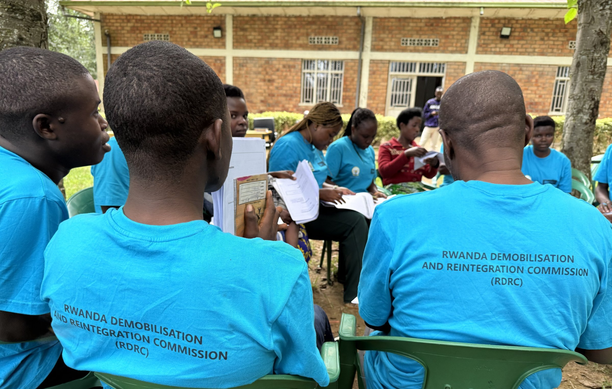 L'image montre un groupe de personnes assises à l'extérieur. Elles portent toutes des t-shirts bleu clair avec le texte "RWANDA DEMOBILISATION AND REINTEGRATION COMMISSION (RDRC)" imprimé au dos. On peut apercevoir des chaises en plastique vertes sur lesquelles elles sont assises. À l'arrière-plan, plusieurs personnes sont en train de discuter, et un bâtiment en briques apparaît légèrement flou derrière eux. L'ambiance semble conviviale et engagée, avec des participants qui semblent suivre une activité ou une formation en lien avec leur programme.