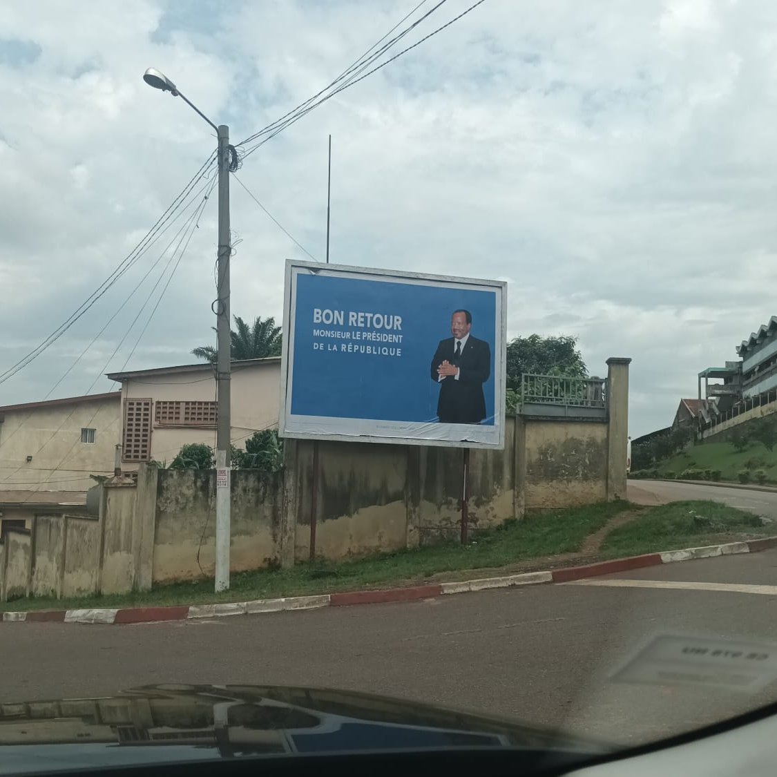 L'image montre une grande affiche publicitaire installée sur un mur à l'extérieur. Sur cette affiche, un homme, probablement un homme politique, se tient debout avec un sourire, vêtu d'un costume. Le fond est bleu, ce qui attire l'attention. En haut de l'affiche, on peut lire le texte "BON RETOUR", suivi d'un message qui semble faire référence à un événement ou une situation spécifique. En dessous, s'étend un paysage urbain avec des bâtiments, des routes et des arbres, et l'image suggère un périphérique ou une route en pente. Le ciel est partiellement nuageux, ajoutant une ambiance calme à la scène.