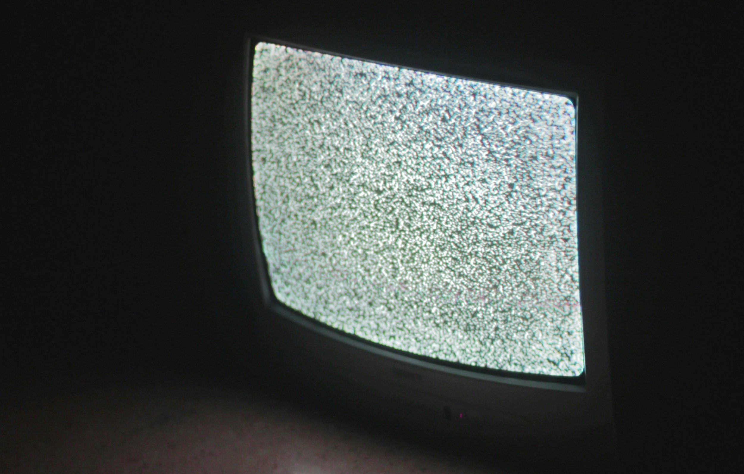 L'image présente un téléviseur ancien, dont l'écran affiche un bruit visuel, souvent appelé "neige", constitué de points blancs et noirs qui scintillent. L'appareil est placé sur le sol, et l'éclairage ambiant est faible, ce qui met en avant le contraste de l'écran lumineux par rapport à l'obscurité environnante. Le téléviseur a des bords arrondis et un design rétro, typique des modèles d'une autre époque. La scène dégage une atmosphère nostalgique et un sentiment d'attente.