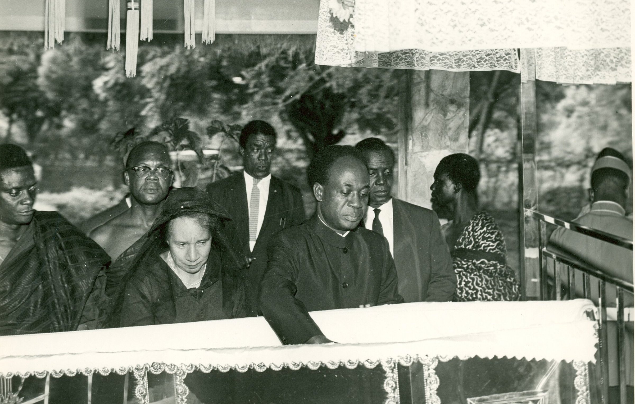 L'image présente une scène de recueillement, probablement lors d'un enterrement ou d'une commémoration. Au premier plan, un homme, habillé en noir, se penche vers un cercueil reposant sur une estrade, symbolisant le respect et la tristesse. À ses côtés se trouve une femme, également en noir, qui semble très émue. En arrière-plan, plusieurs personnes regardent la scène avec un air sérieux, toutes vêtues de manière formelle, ce qui souligne le caractère solennel de l'événement. L'atmosphère est empreinte de respect et de recueillement, avec une ambiance calme, interrompue seulement par les murmures des participants.